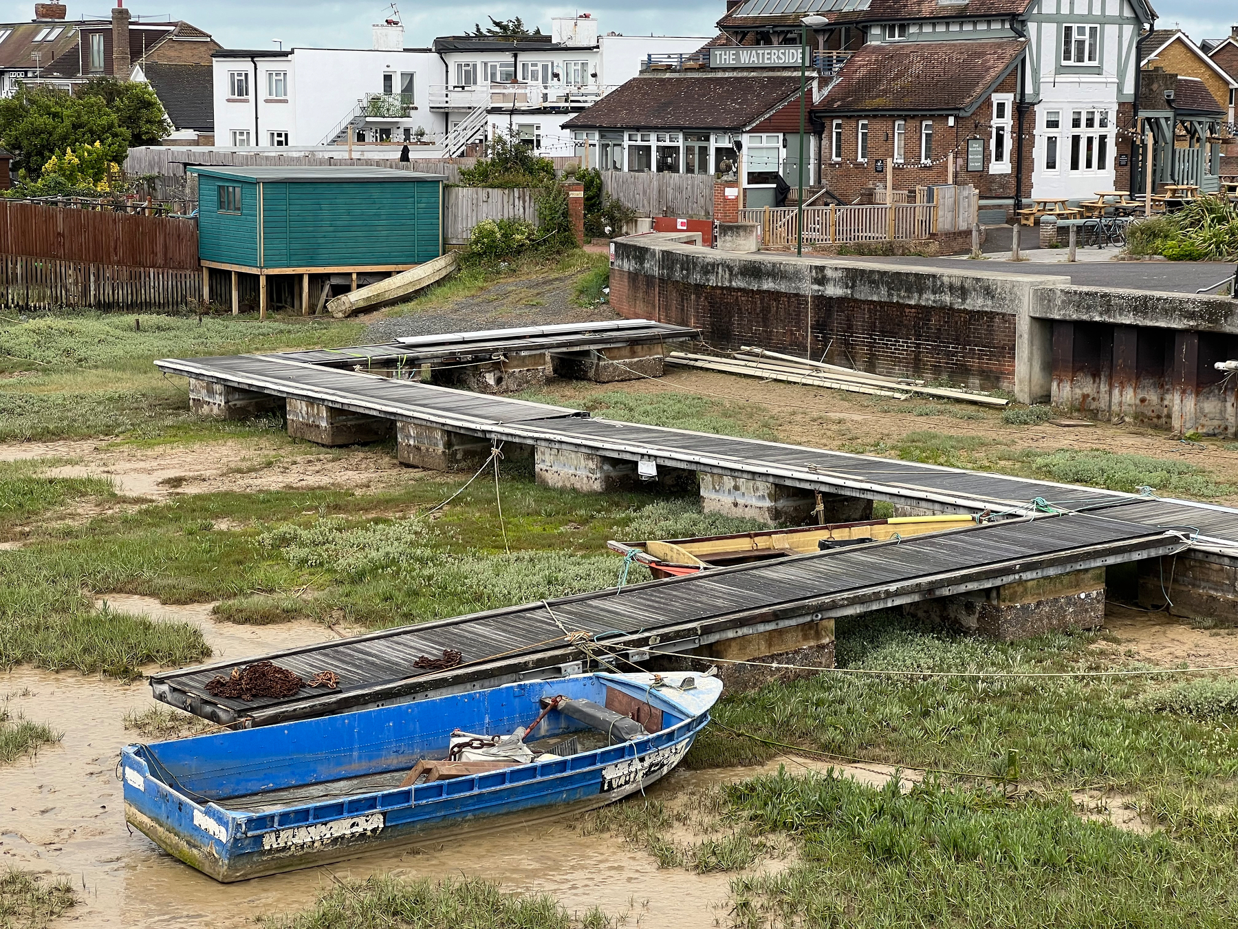 A small blue boat is grounded on a muddy shore near a series of wooden docks, with houses, a green shed, and a building labeled The Waterside in the background.
