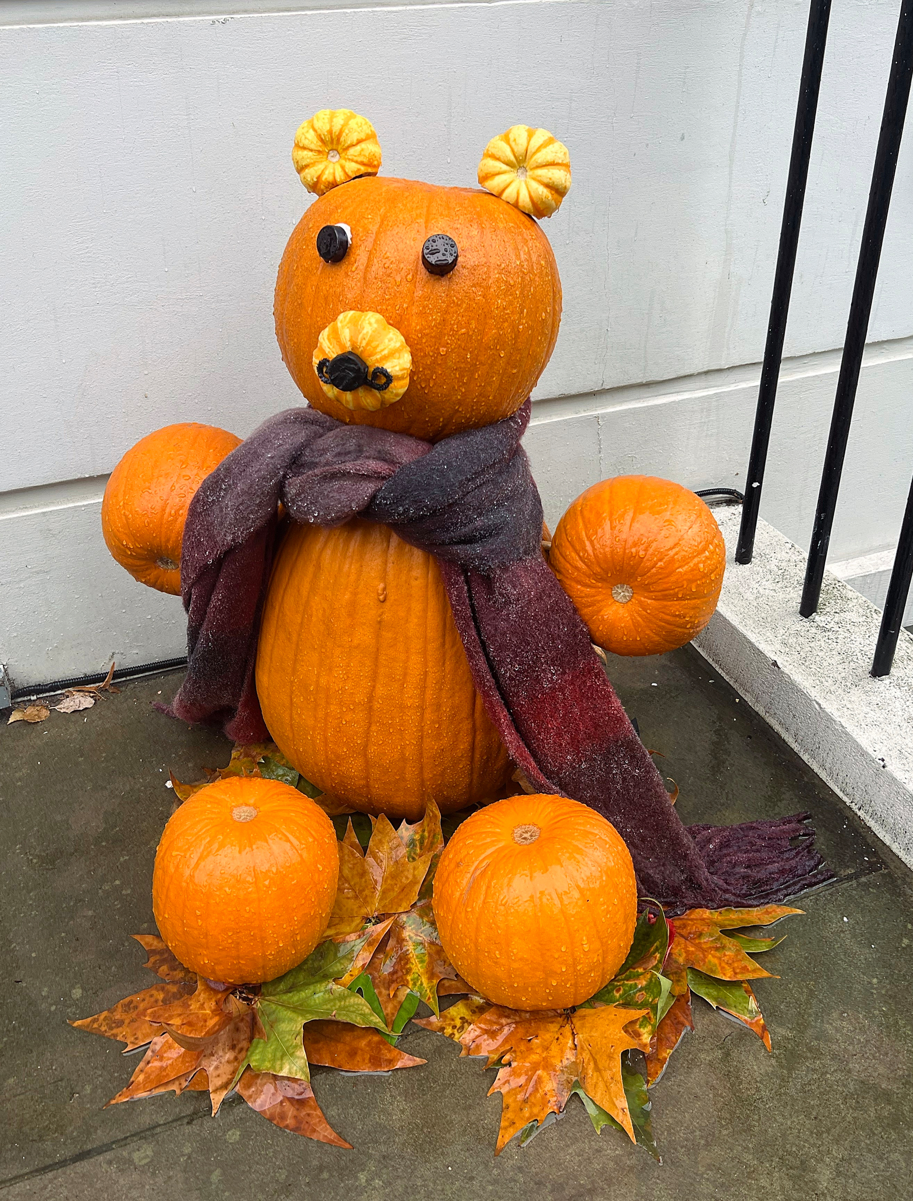 A bear figurine made of pumpkins, with a scarf wrapped around its neck, is sitting on a bed of autumn leaves.