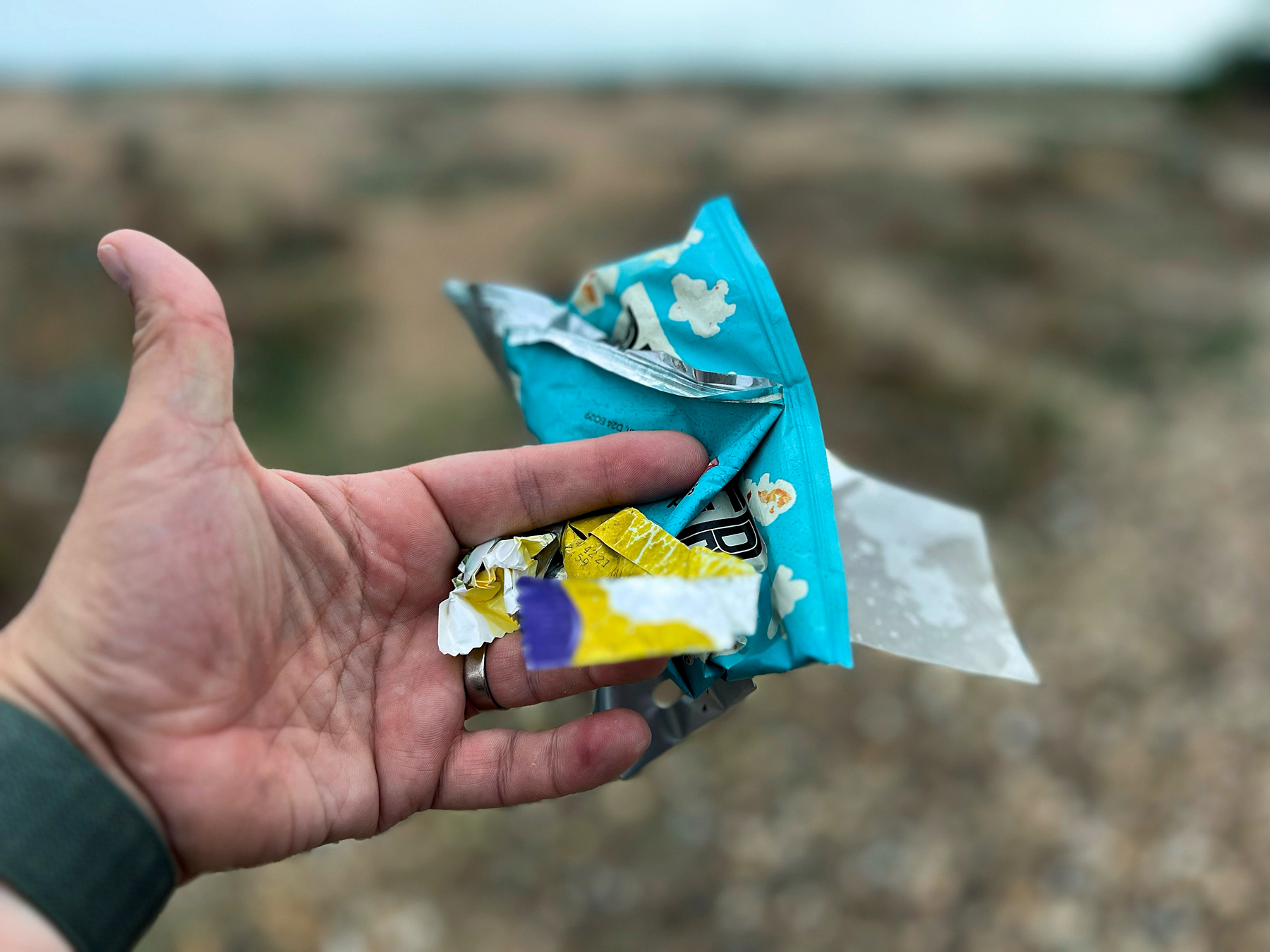 A hand is holding several pieces of crumpled and torn food packaging in an outdoor setting.