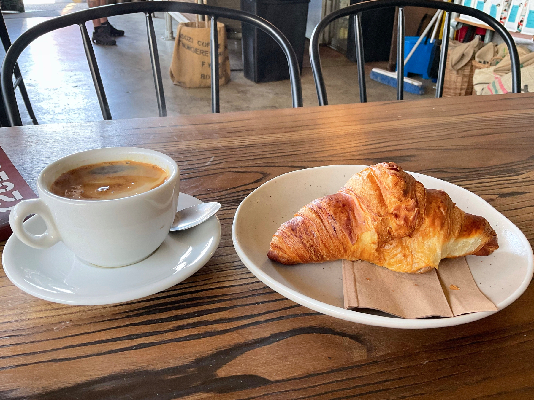 A cup of coffee and a vegan croissant sit on a wooden table at a café.