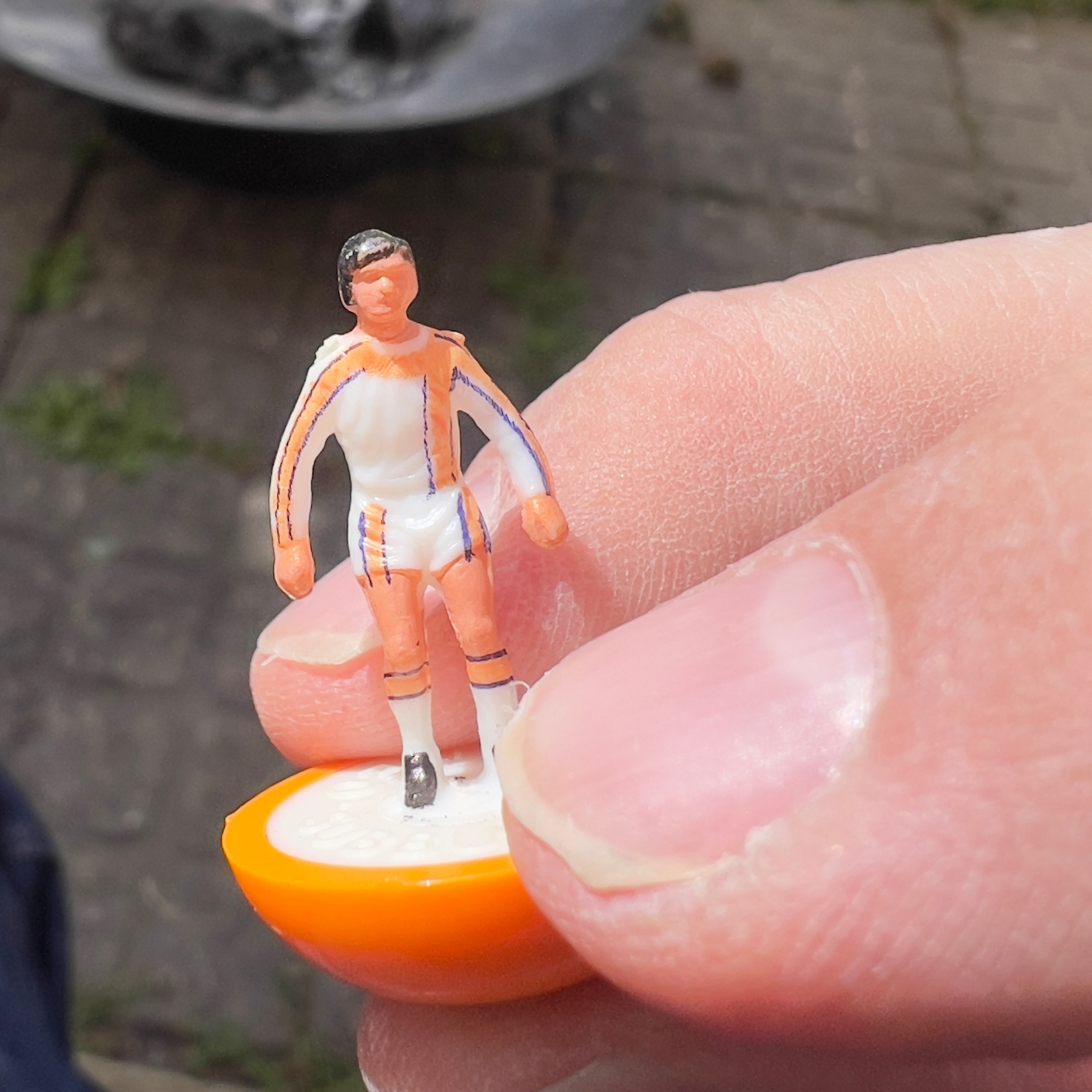 A small figurine of a Subbuteo soccer player, painted in orange and white, is being held between two fingers.