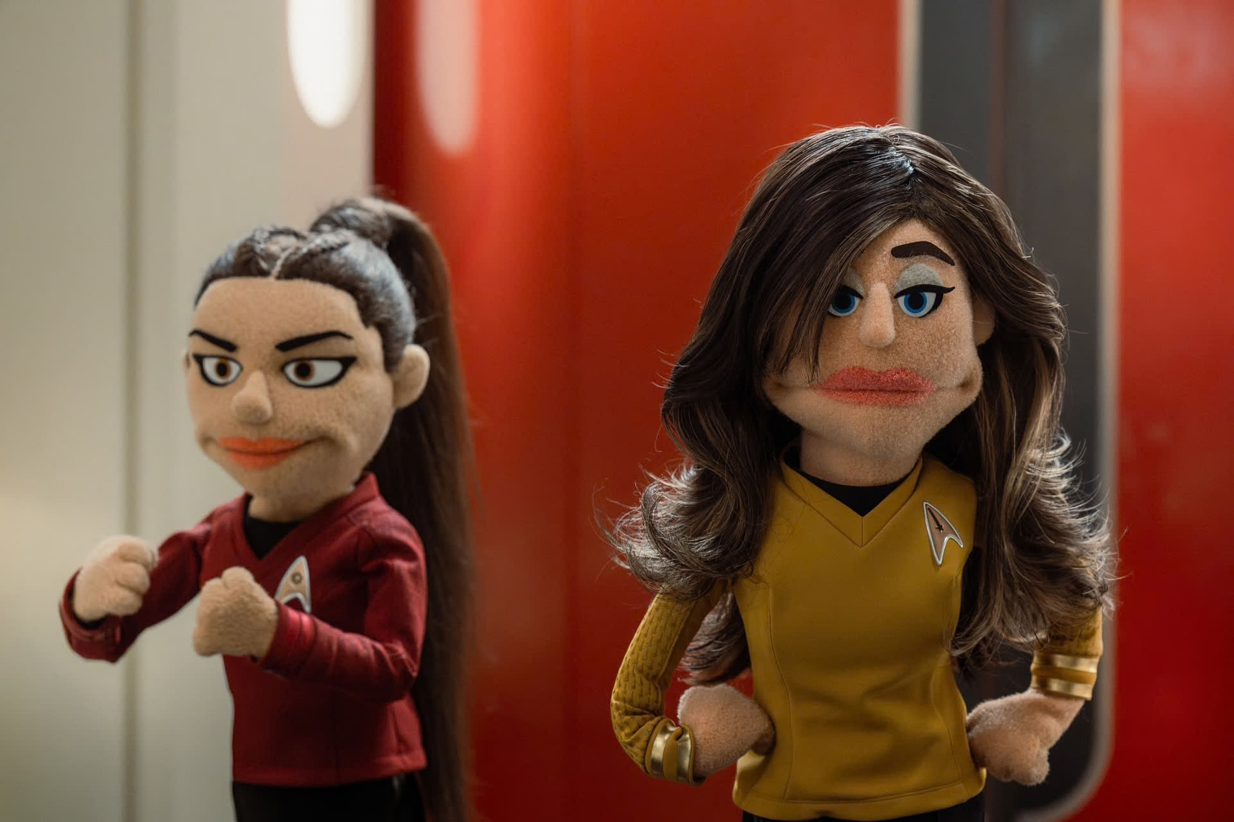 Two puppets dressed in Star Trek uniforms stand in a futuristic setting with one in a red shirt and the other in a yellow shirt.