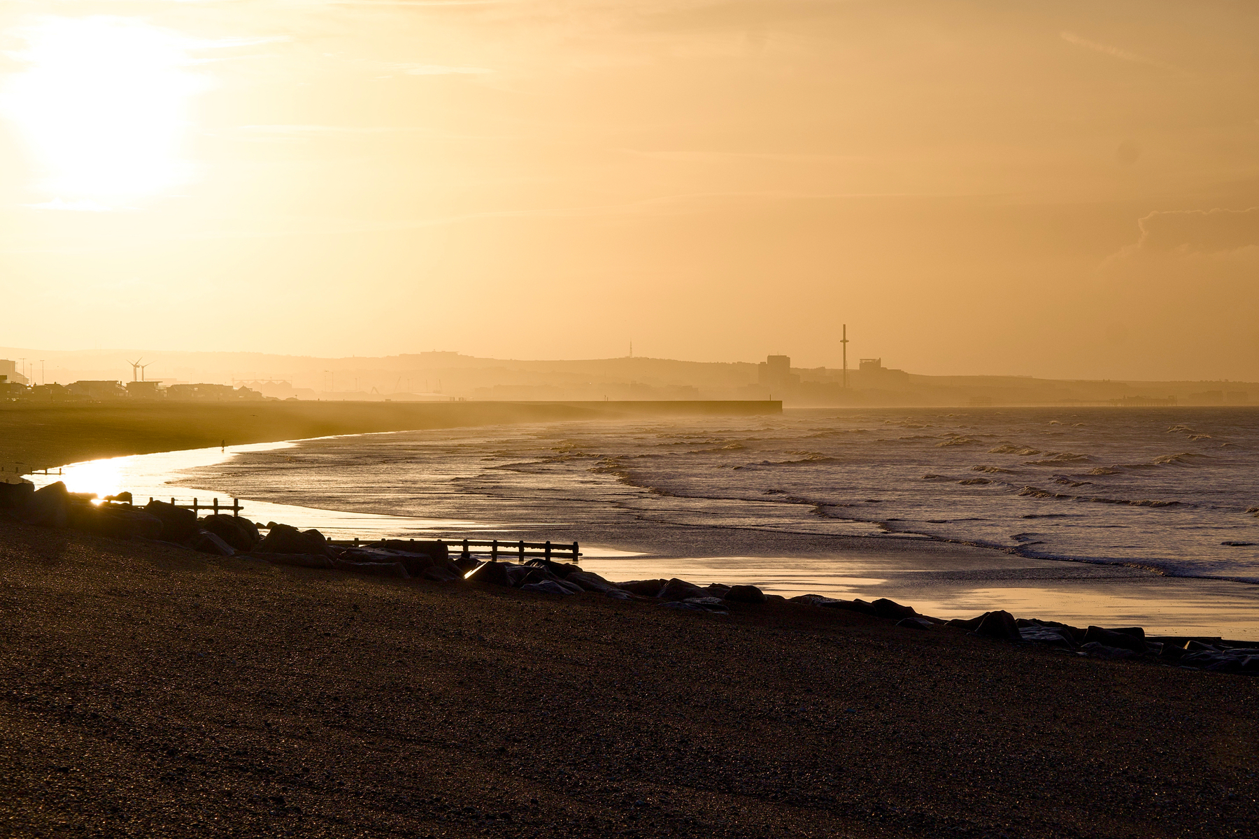 A serene coastal scene at sunrise with gentle waves lapping the shore and a silhouette of distant buildings on the horizon.
