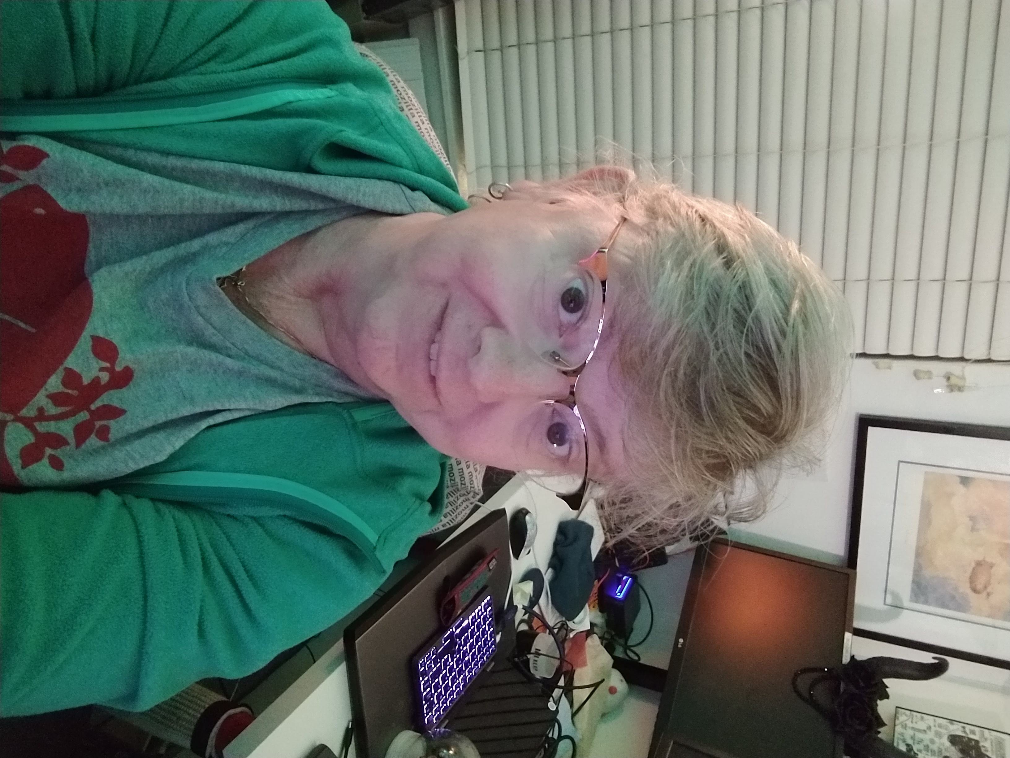 A woman in a grey Roots Canada tshirt and a green hoodie looks up at the camera from her desk. In the background are several computers.