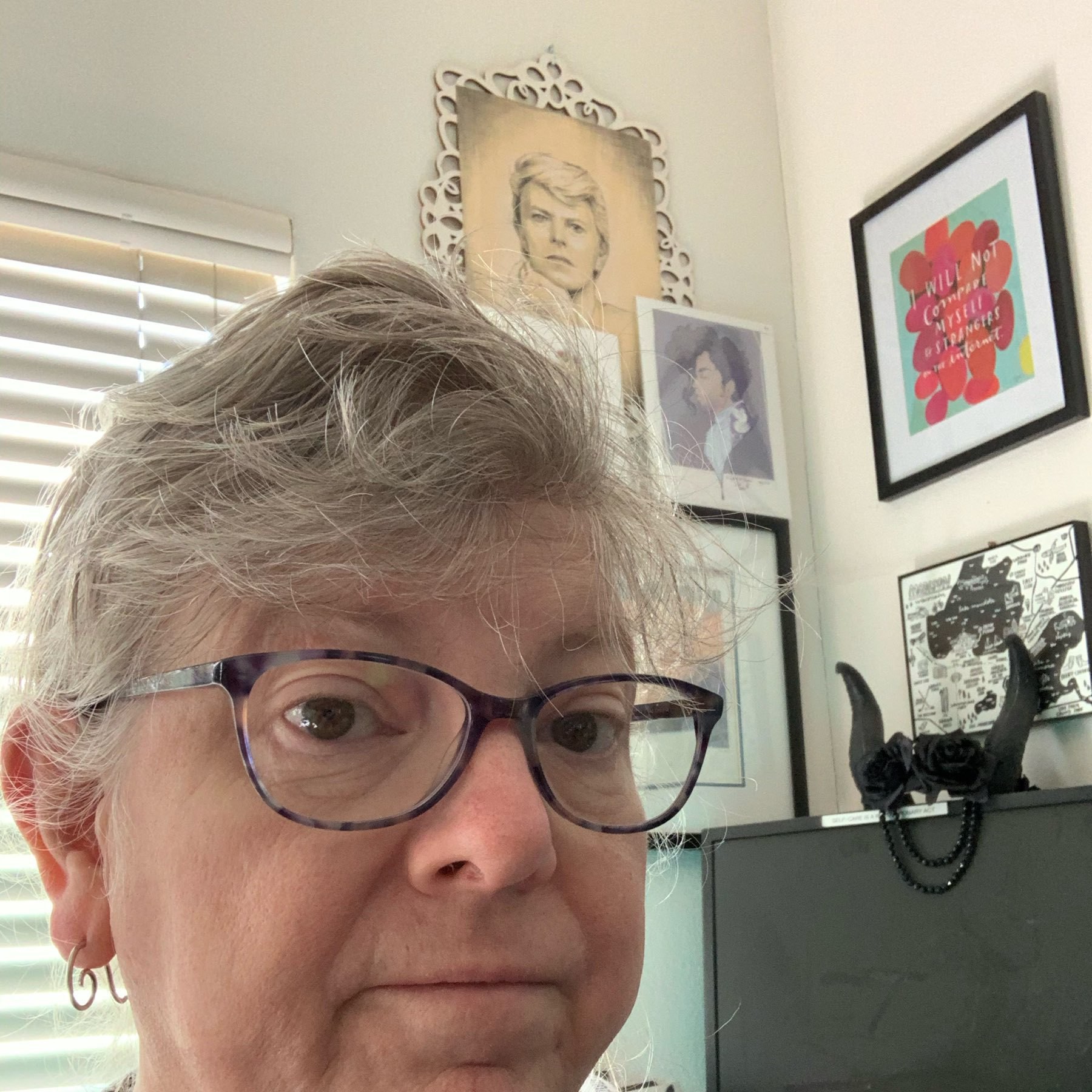 head of a tired white blonde non-binary woman, hair up, thier desk in the background, portraits of bowie and prince above the desk