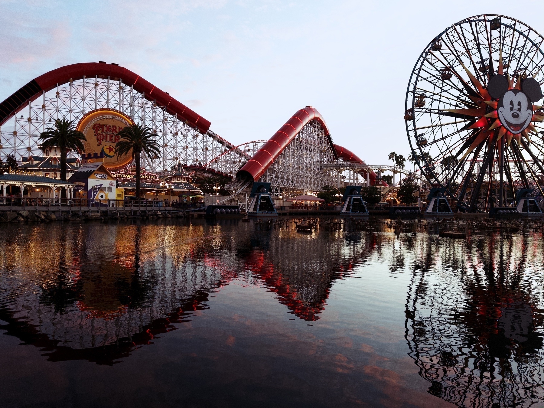 Pixar Pier in the golden hour. The Incredicoaster and Fun Wheel reflect on the pool in the foreground.