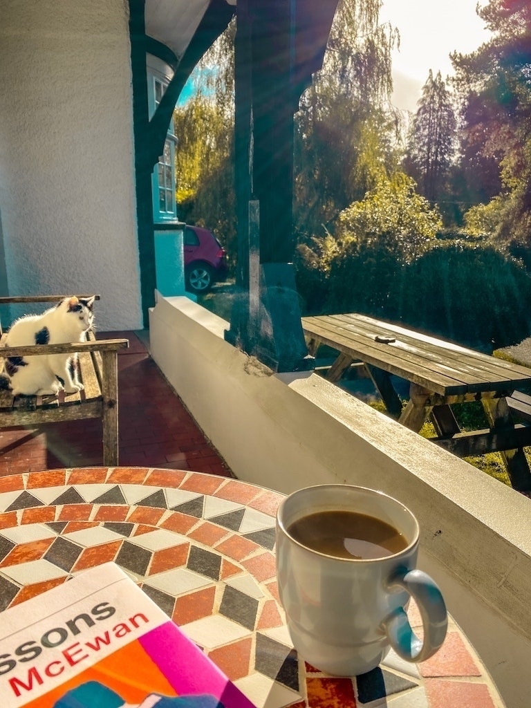 Mug of coffee and book on a mosaic table, cat in the background, sun shining
