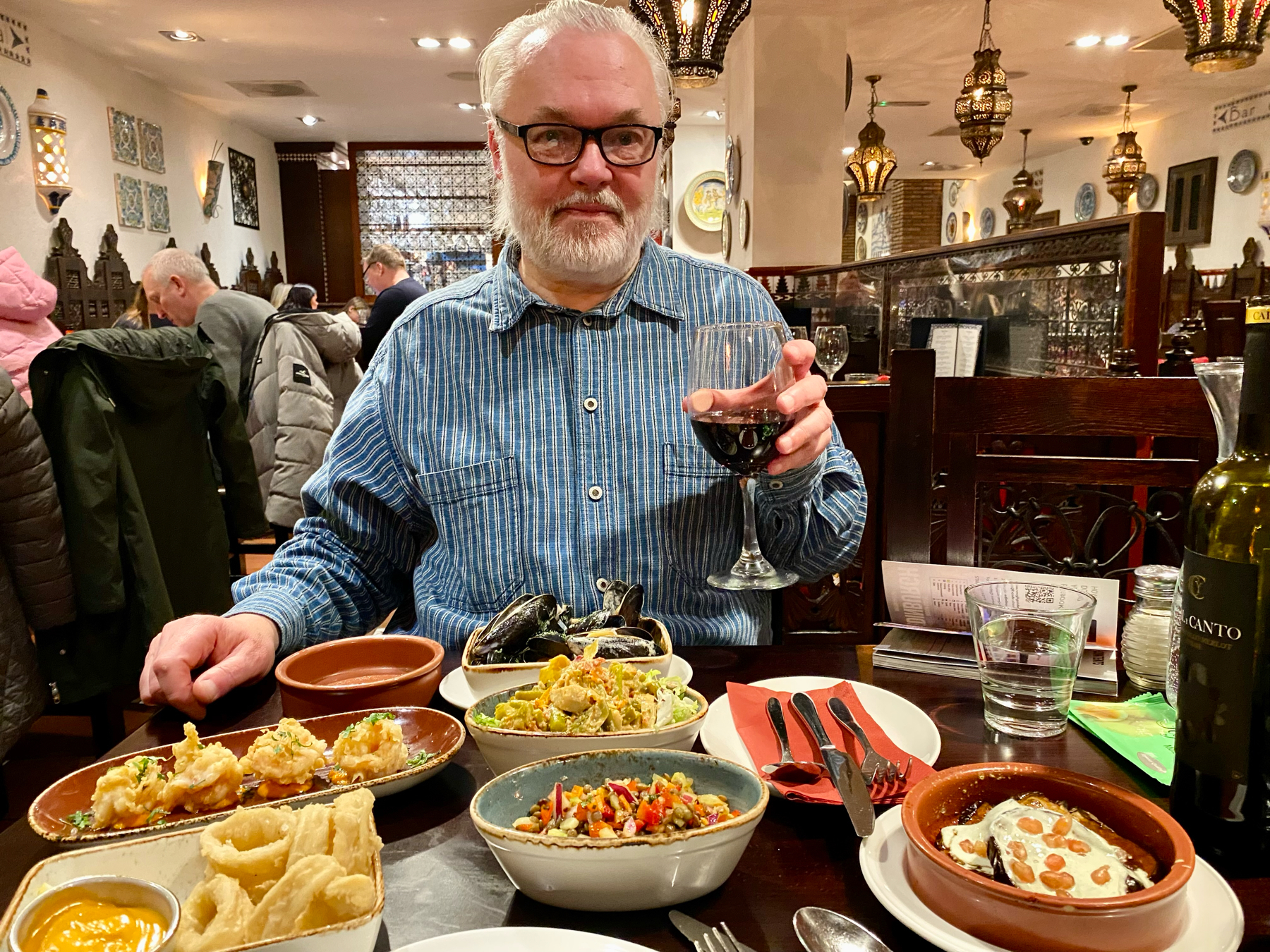 Man holding a glass of wine at a table covered with small plates of food