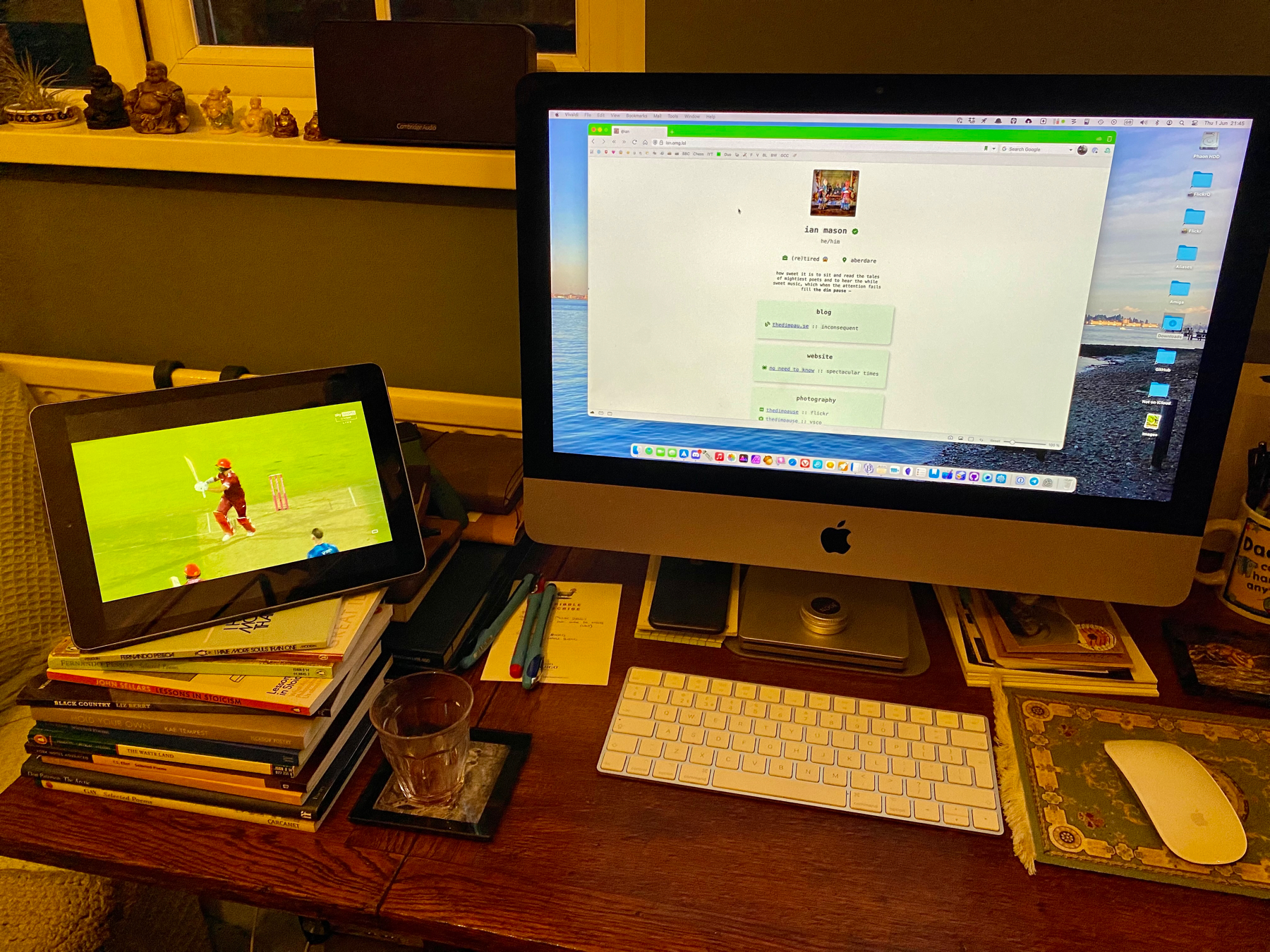 iPad showing live cricket match propped up next to an iMac, both on a crowded, messy desk