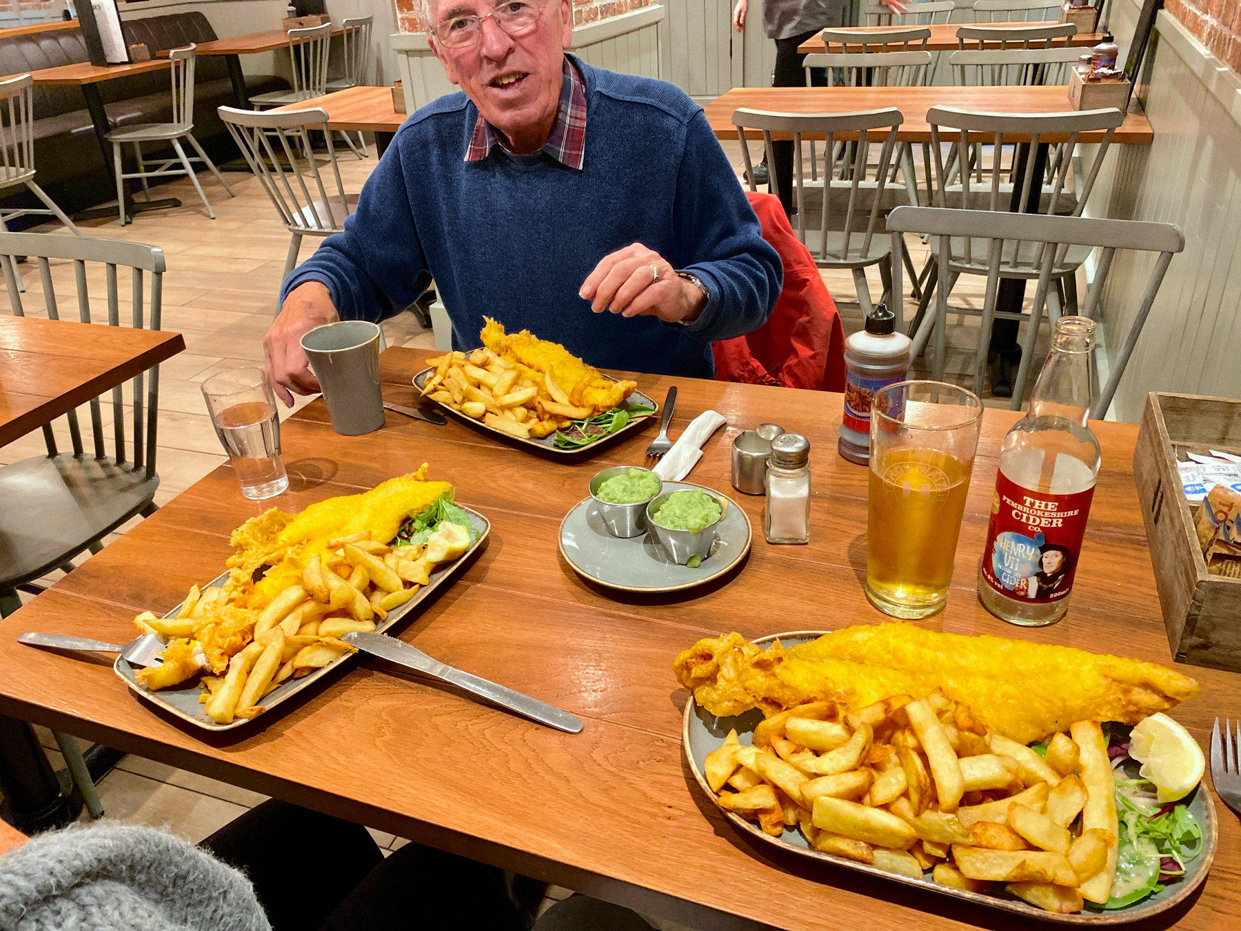 Cafe table with three plates of fish and chips and drinks, man sitting at far side of table