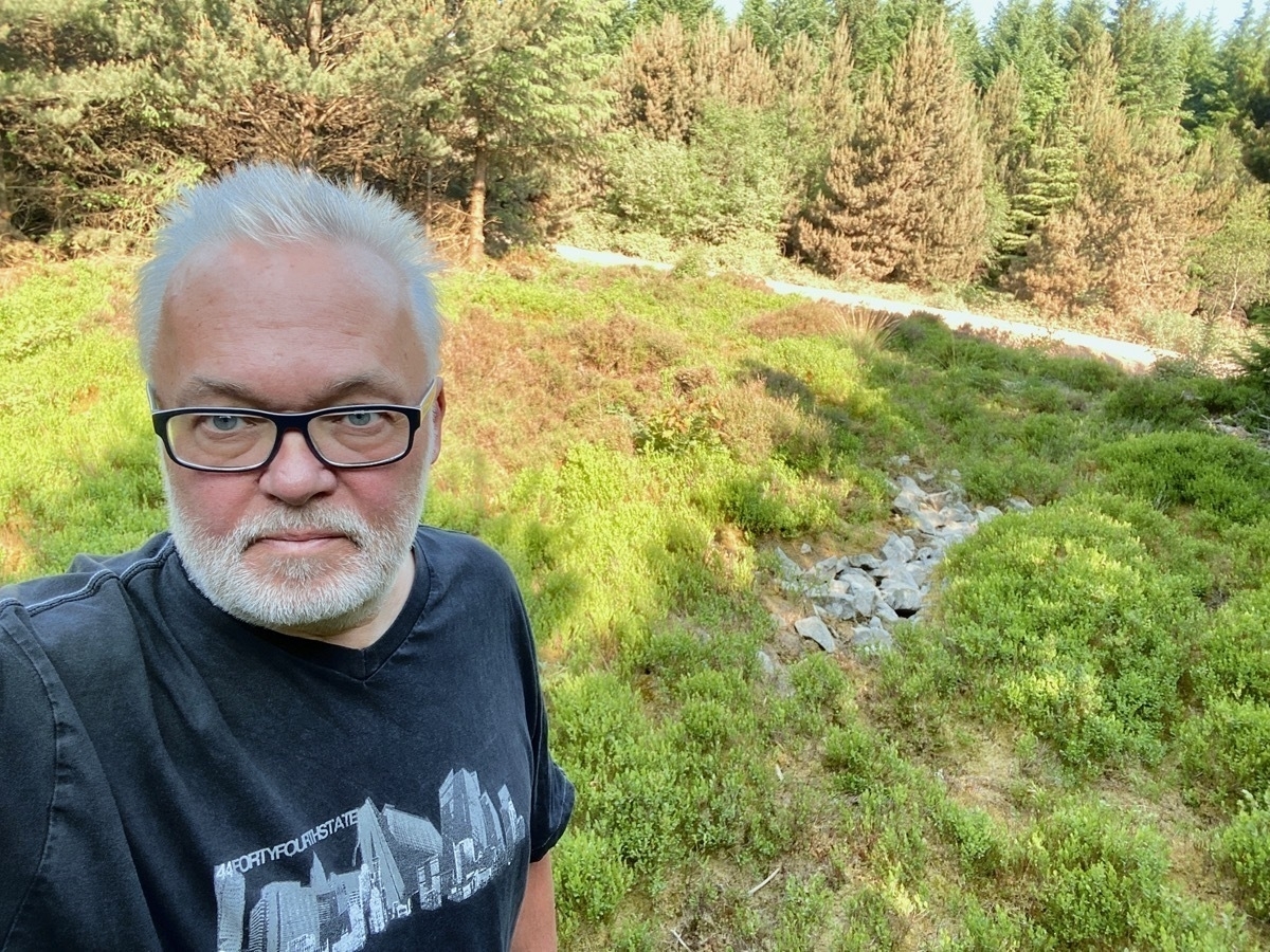 Selfie of bearded man in on scrub ground with small area of stones visible, pine trees in background
