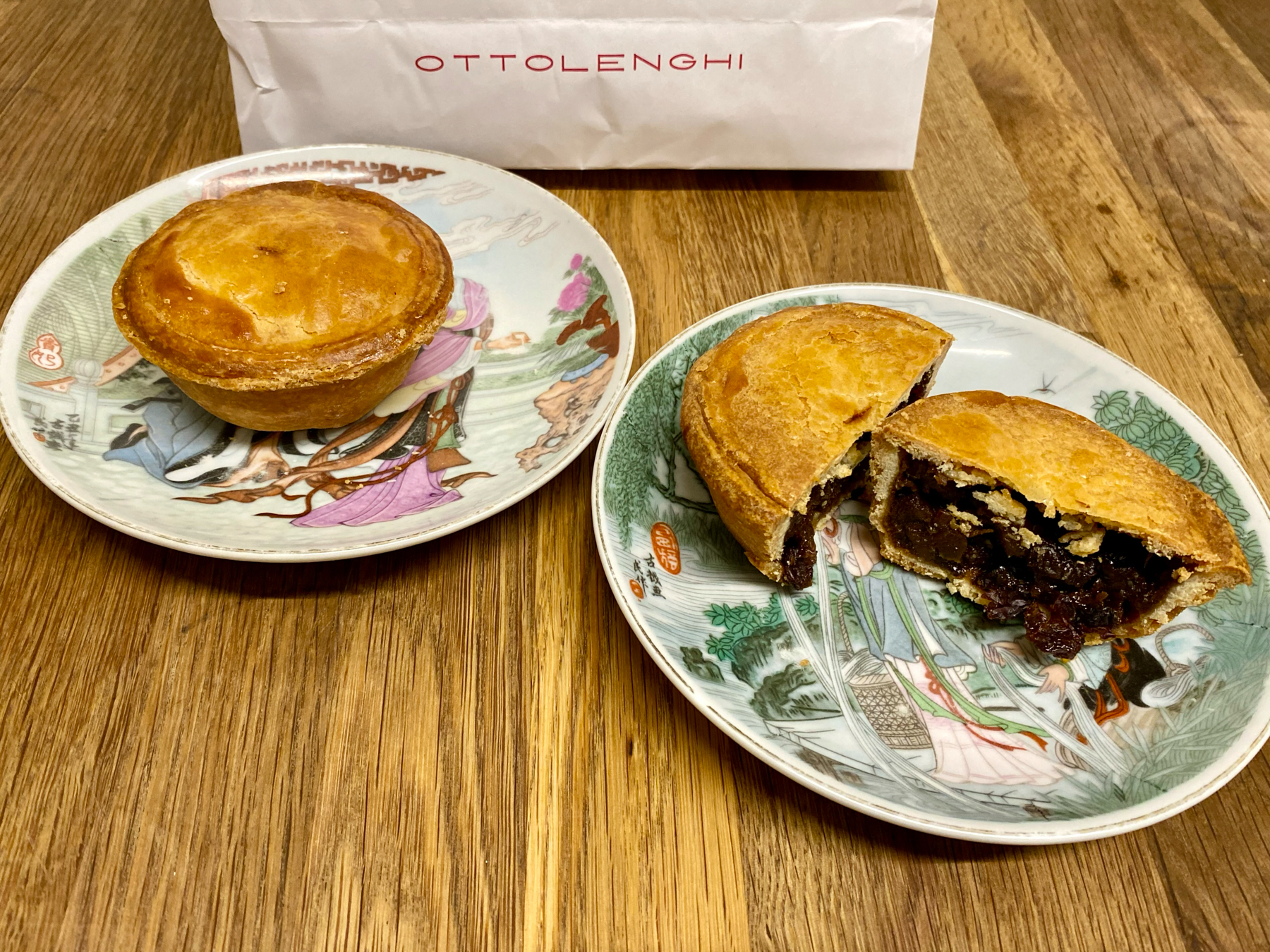 A whole pie and a sliced open pie revealing a dark filling on decorative plates, with a OTTOLENGHI branded bag in the background, all on a wooden surface.