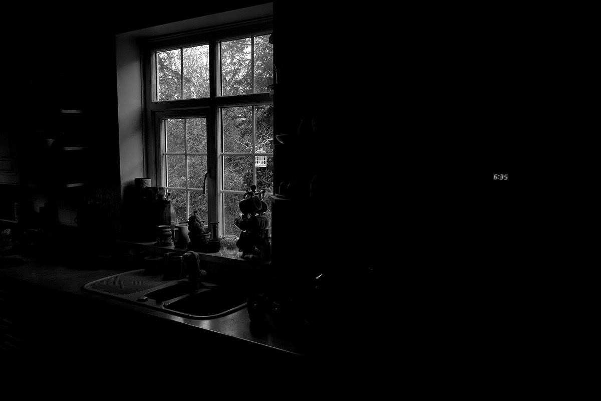 Monochrome image of darkened kitchen window, digital clock visible to left showing 6:35