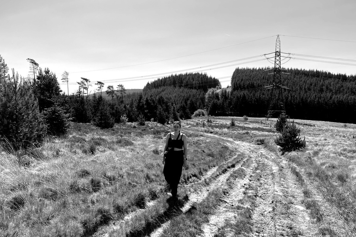 Monochrome image of woman walking on path through field, backdrop of conifers and electricity pylon