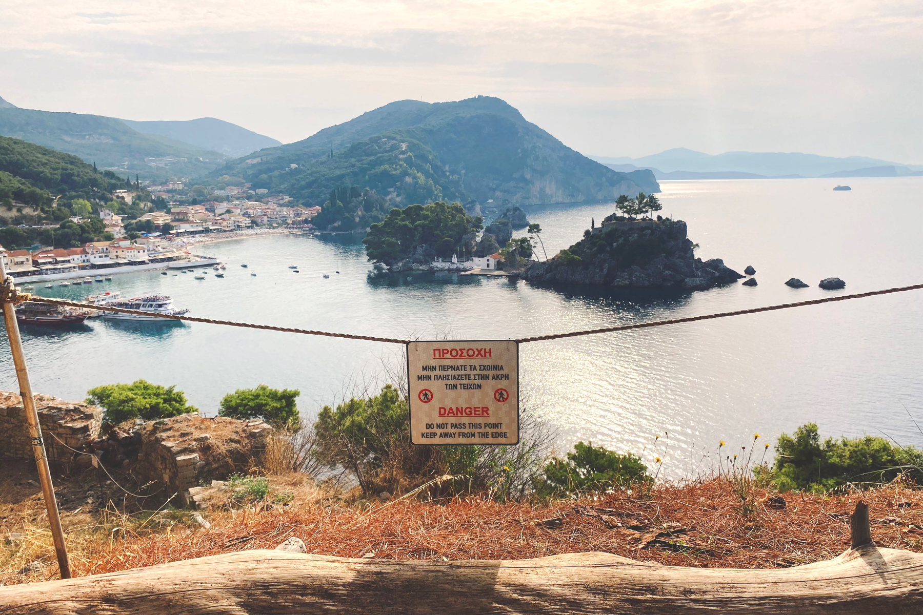 View from the edge of a cliff overlooking the sea, a small town and harbour and several small islands. A danger sign is suspended on a rope in the foreground warning about keeping away from the edge