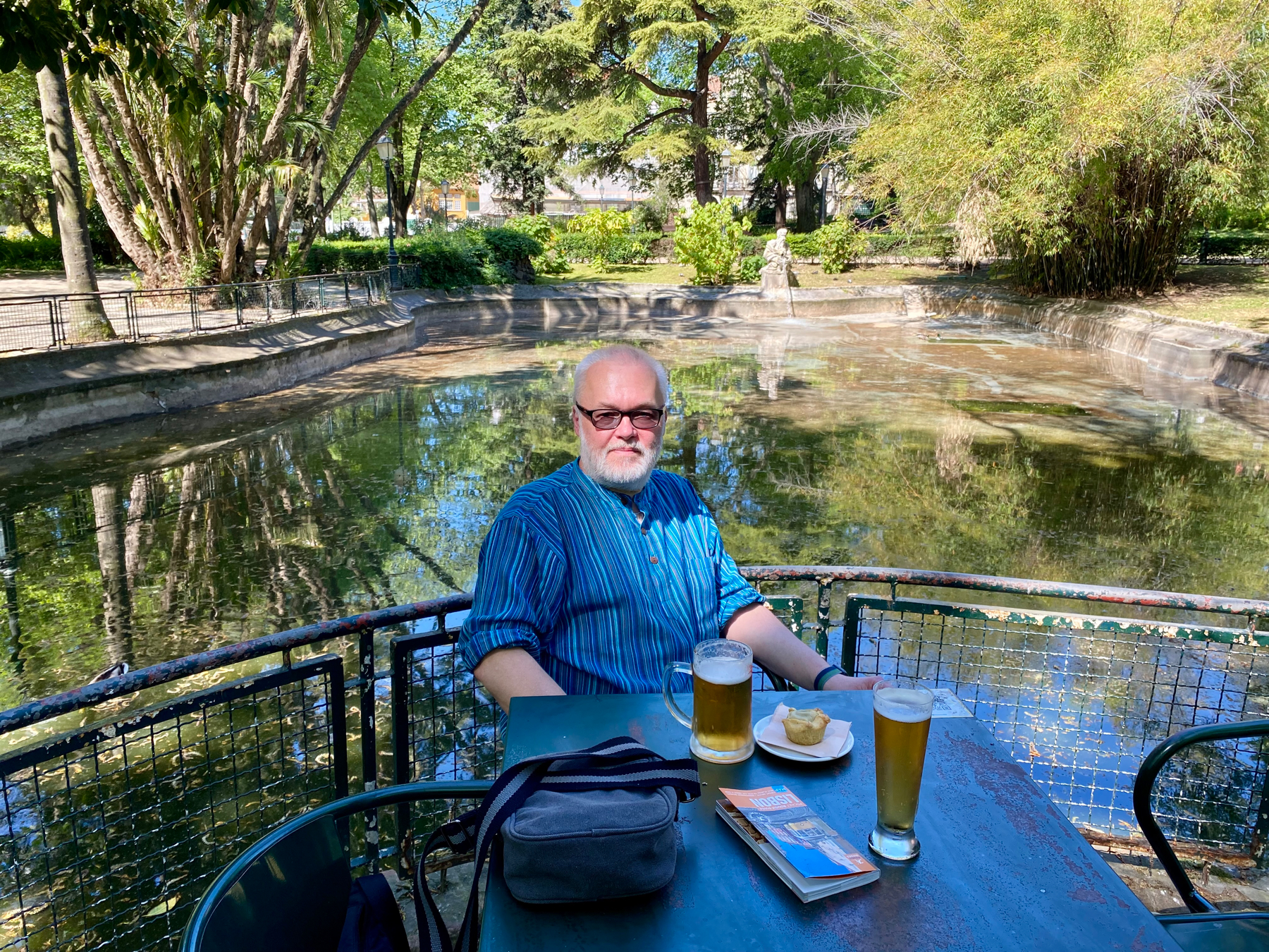 Man seated at a table with beers and a small pie. In a park, pond in background