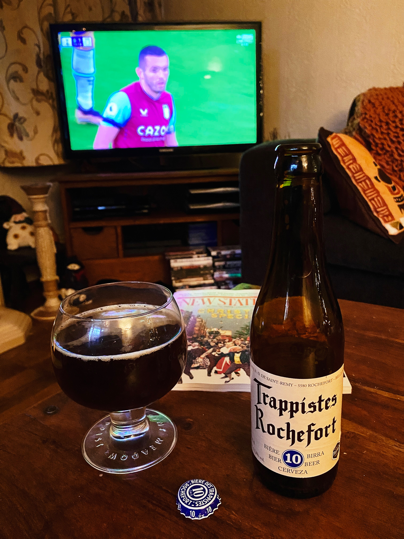 Bottle and glass of dark Belgian beer, football match on TV in the background