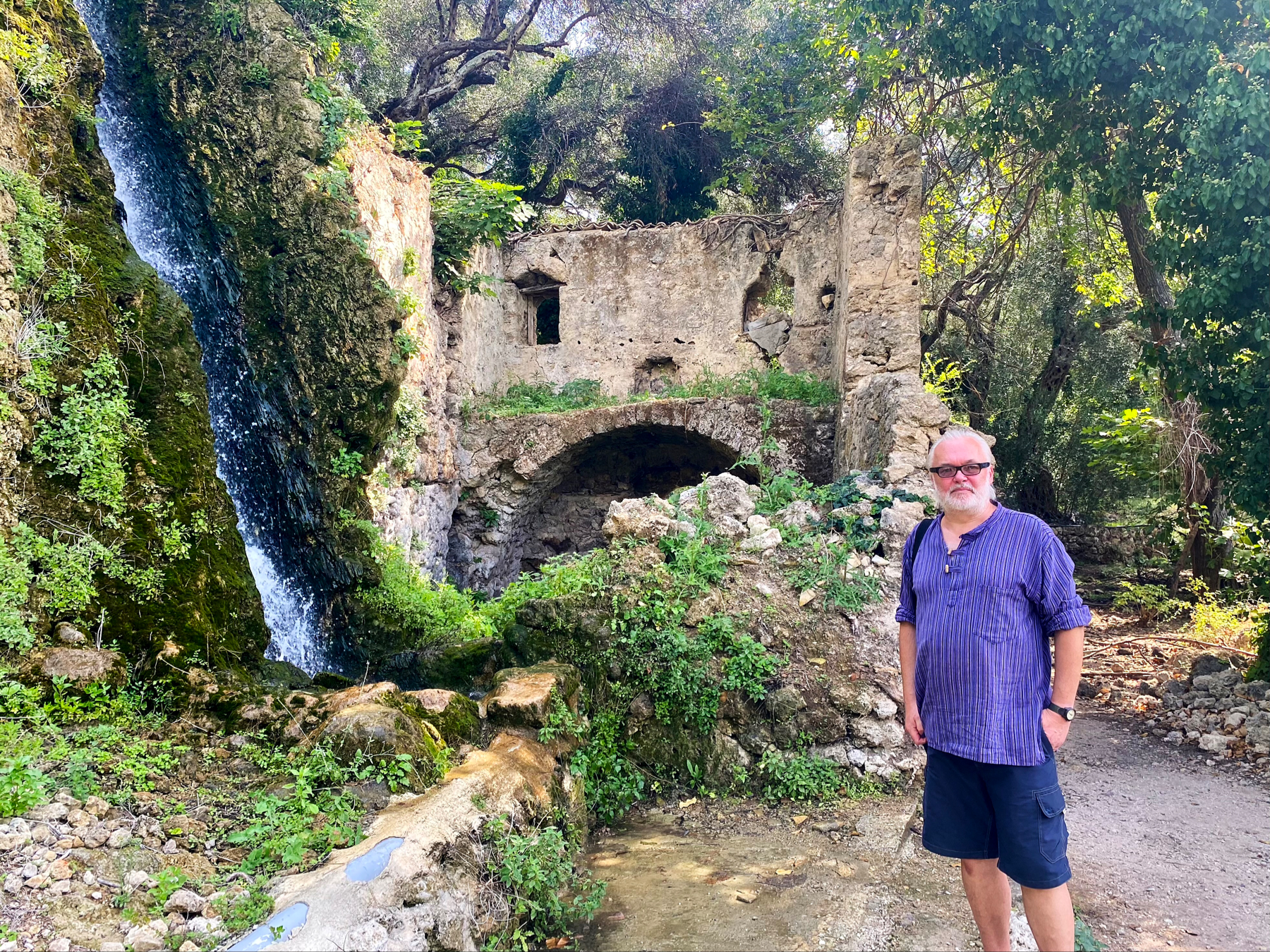 Man standing next to foliage covered ruined building. Stone archway and vertical cascade of water visible 