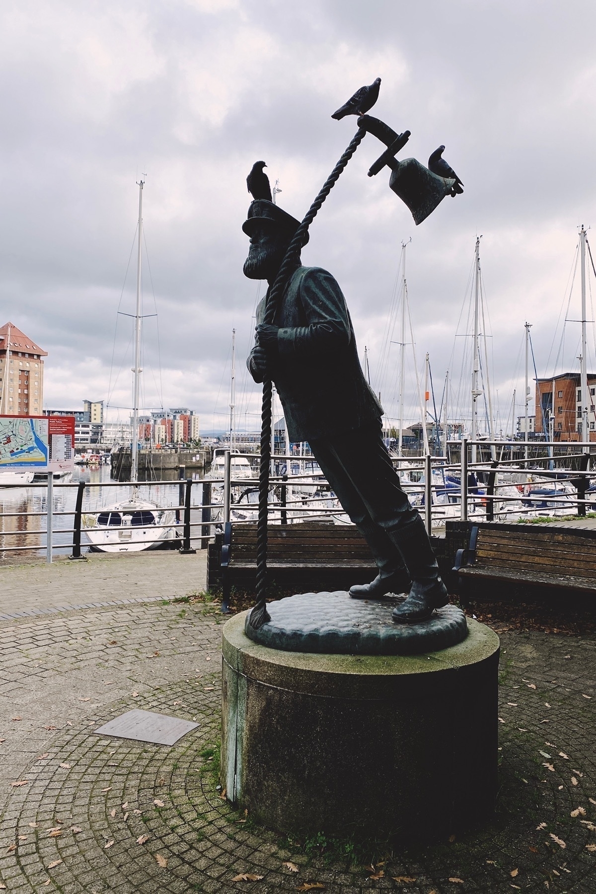 Statue of a sea captain leaning forward grasping a rope with bell attached. Three pigeond perched on statue. Moored boats in background.