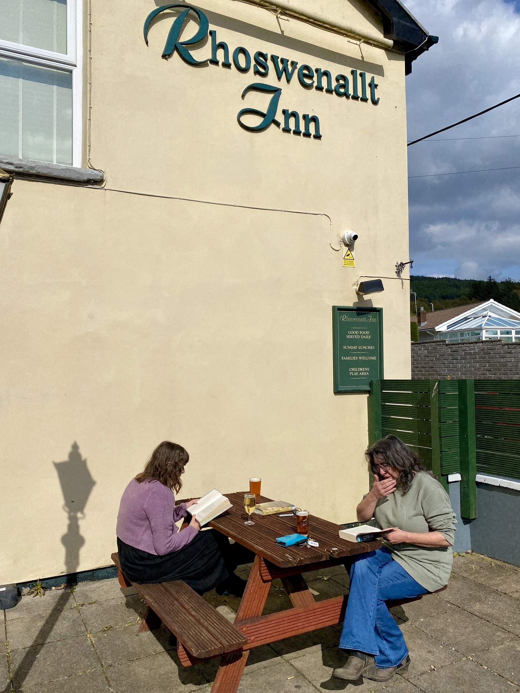 Two women reading books on a picnic bench outside a pub. Glasses of beer on the table.