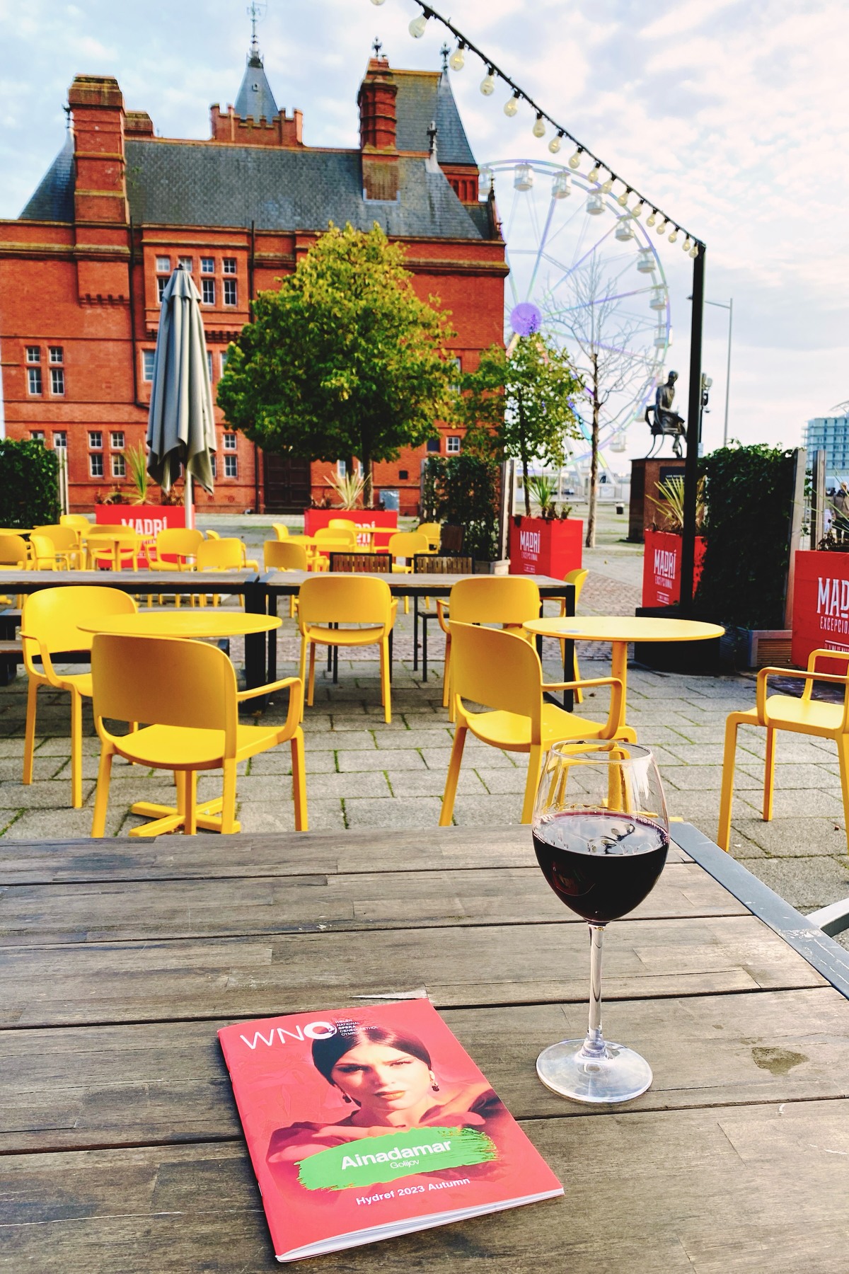 Glass of red wine next to a booklet on an outdoor table with yellow chairs, a red brick building and Ferris wheel in the background 