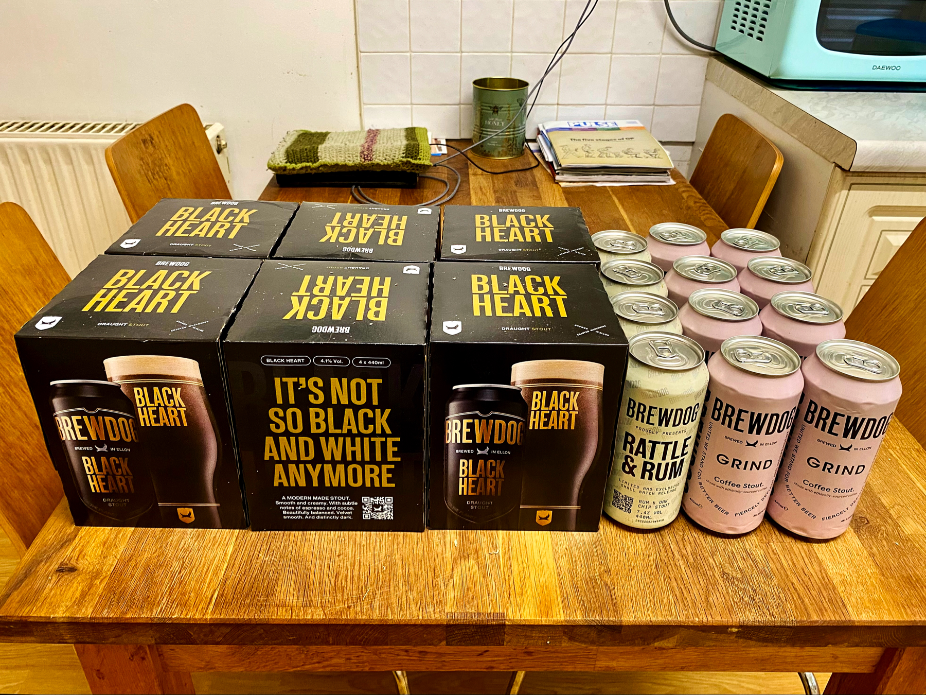 Boxed packs and cans of beer on a wooden table. Boxes show a picture of a glass of dark beer, cans are black, cream and pink