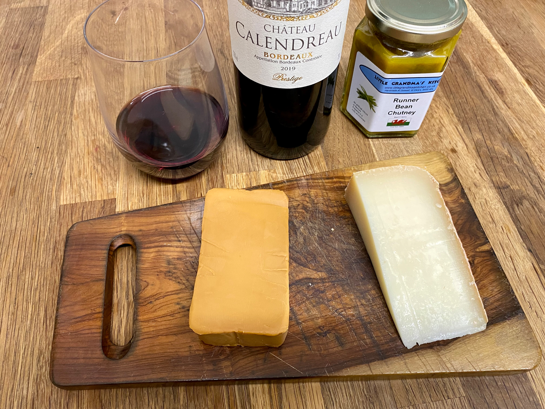 Two blocks of cheese on a wooden board, one of them a caramel brown colour. Red wine and chutney in the background.