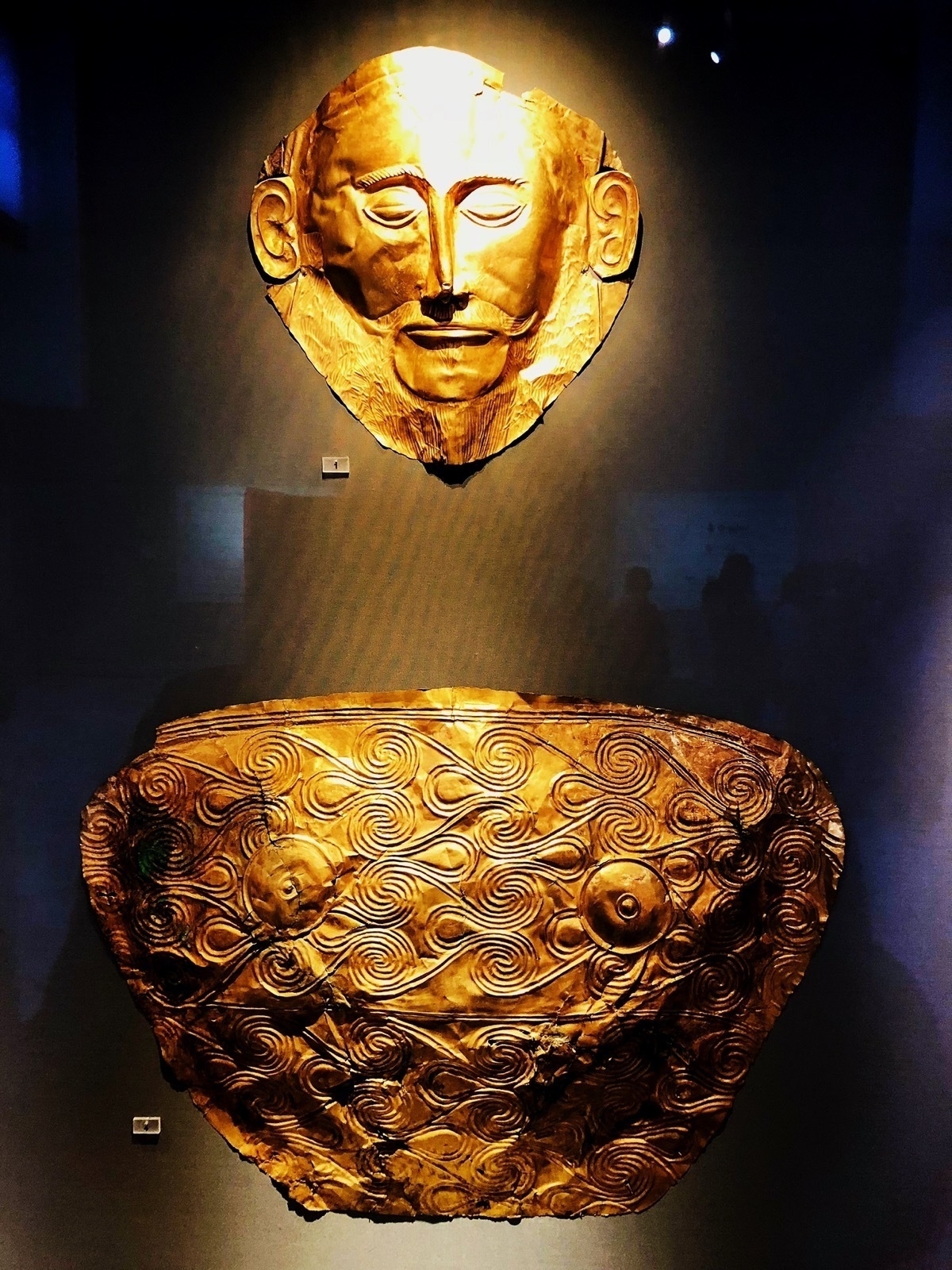 Shining gold mask of a bearded man above a decorative gold sheet chest piece, both mounted on a dark background