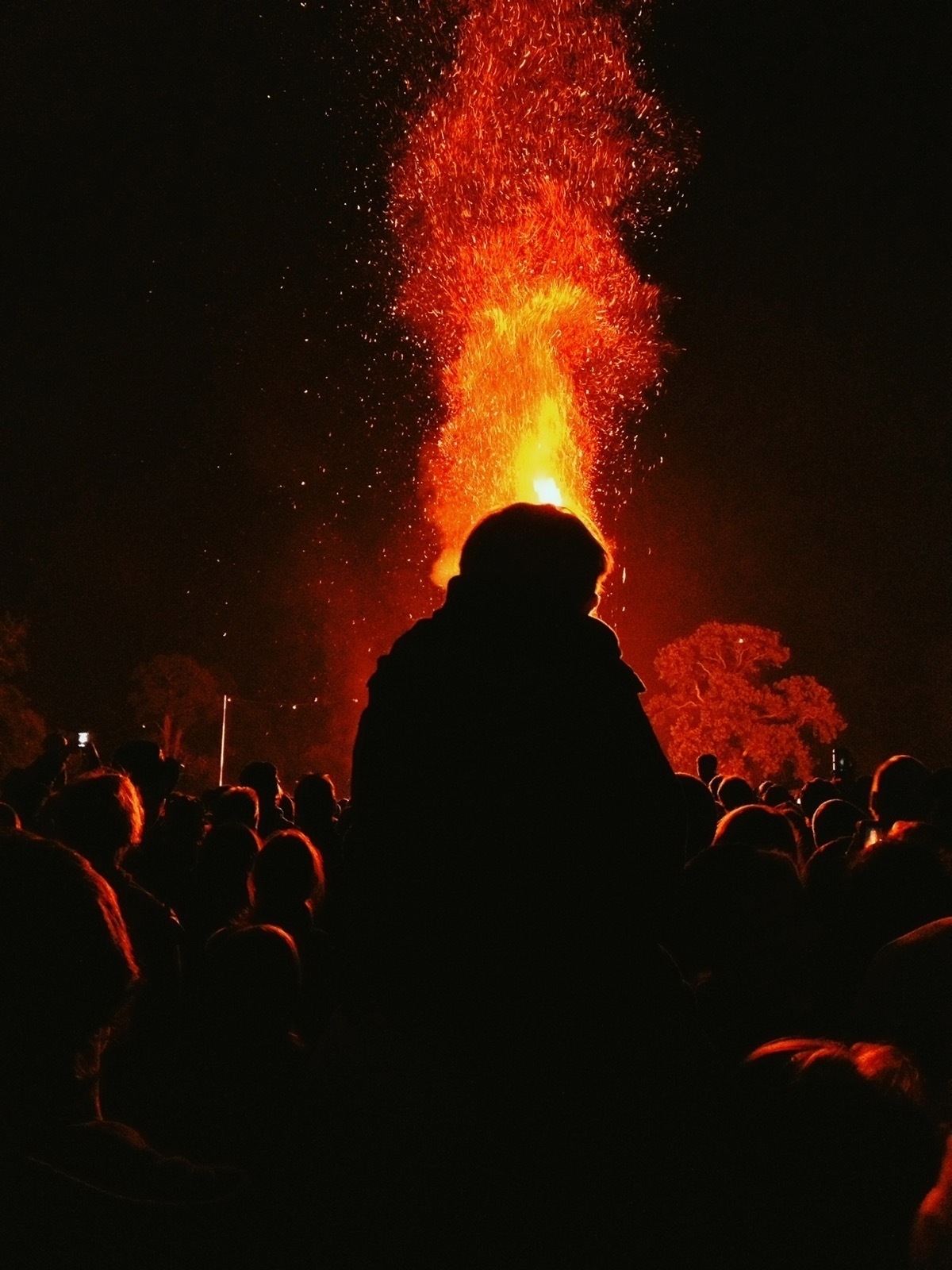 Silhouette of a boy on a man's shoulders above a crowd of people, in front of a plume of sparks from a bonfire