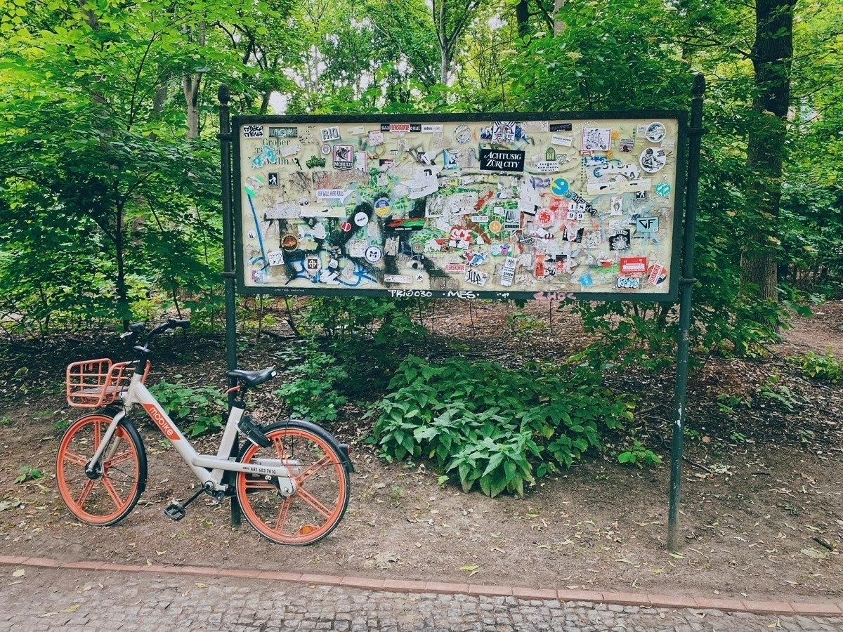 Orange and silver e-bike leaning against a large noticeboard covered in stickers. Foliage in background.