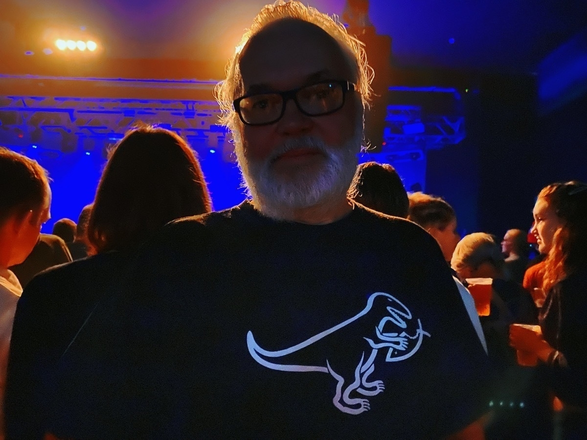 Man with grey hair, beard and glasses in a crowd inside a concert venue