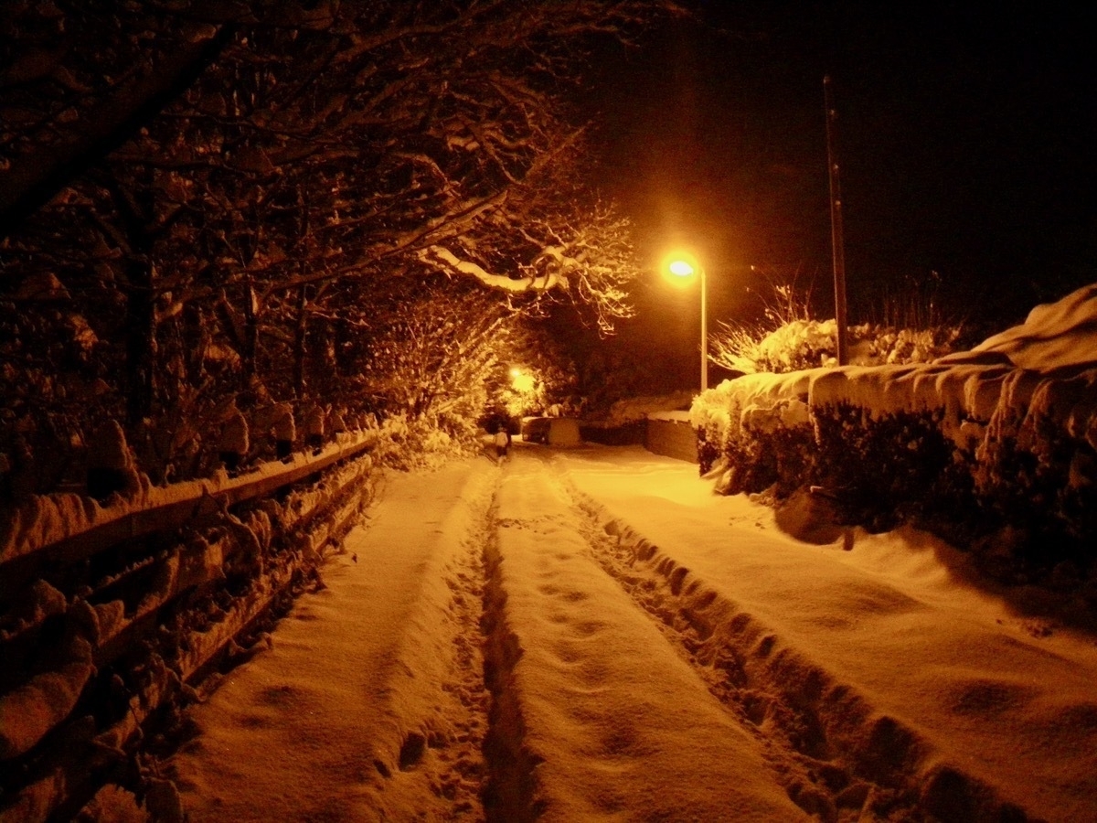 Streetlight glowing at night - orange illumination over a country lane which is several inches deep in snow. Deep tyre tracks in the snow.