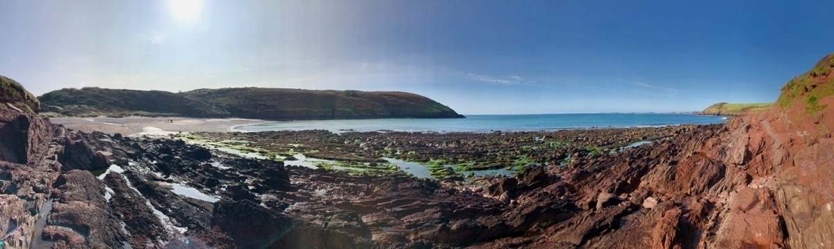 Panoramic view of a small bay. Rocks and rock pools in foreground, small sandy beach and low cliffs and hills in background. Blue skies and sun.