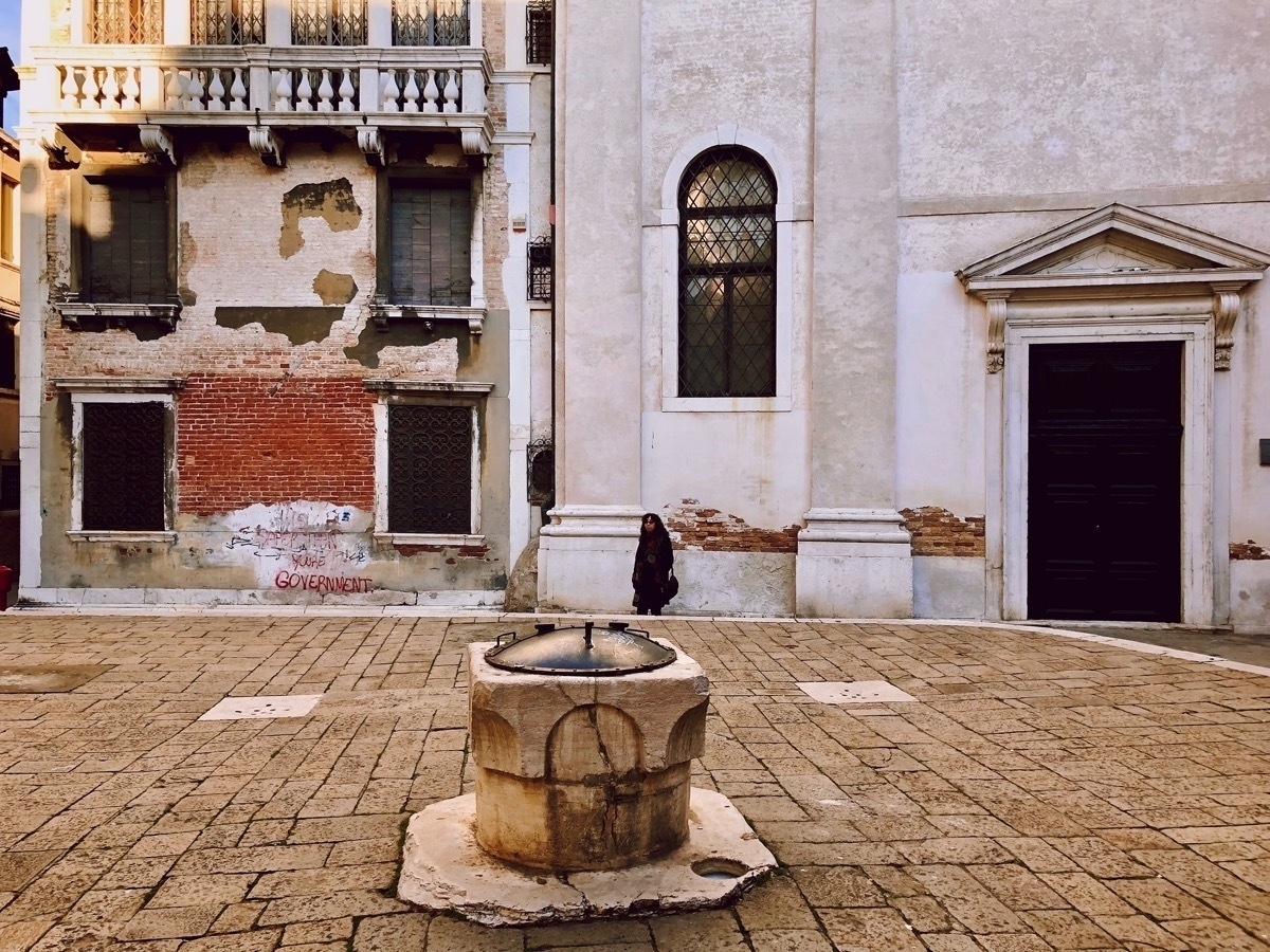 Stone well, capped with a metal dome, on a stone paving floor in a Venetian square with old church building and brick wall in the background