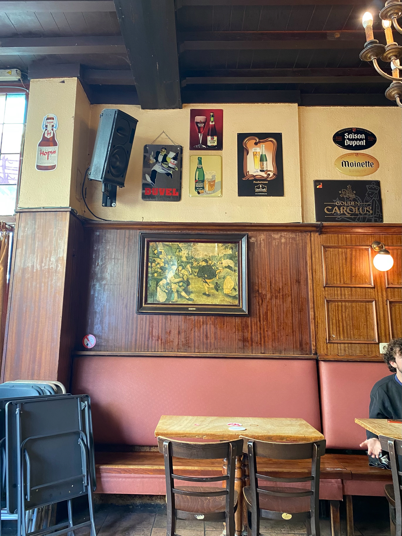 A cozy pub interior with wooden elements and red cushioned seating. The walls display various vintage beer advertisements, including brands like Duvel, Saison Dupont, Moinette and Gouden Carolus. A framed painting hangs above  a small table.