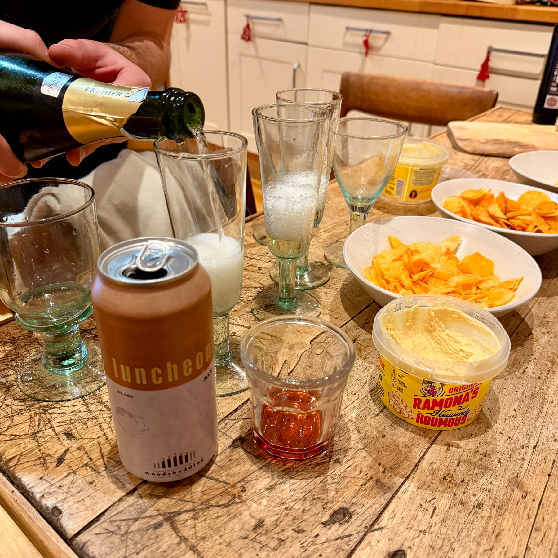  A person is pouring champagne into glasses on a table with snack bowls, hummus, and a drink can.