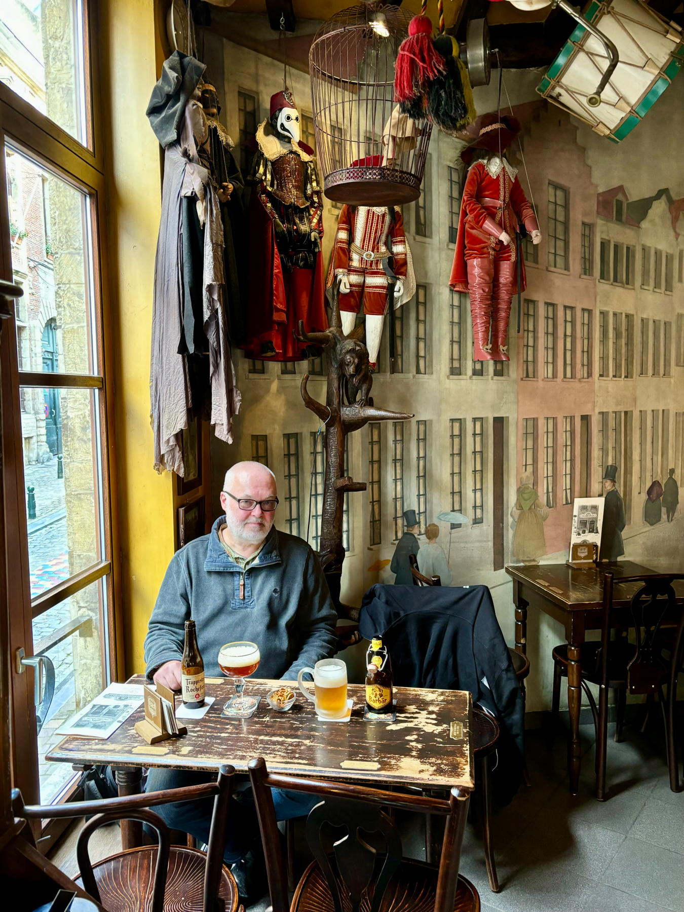 A man sits at a rustic wooden table in a cozy café, drinking beer. The table has two bottles of beer and a bowl of snacks. The interior decor features hanging vintage marionette dolls and a mural of old European buildings and people.