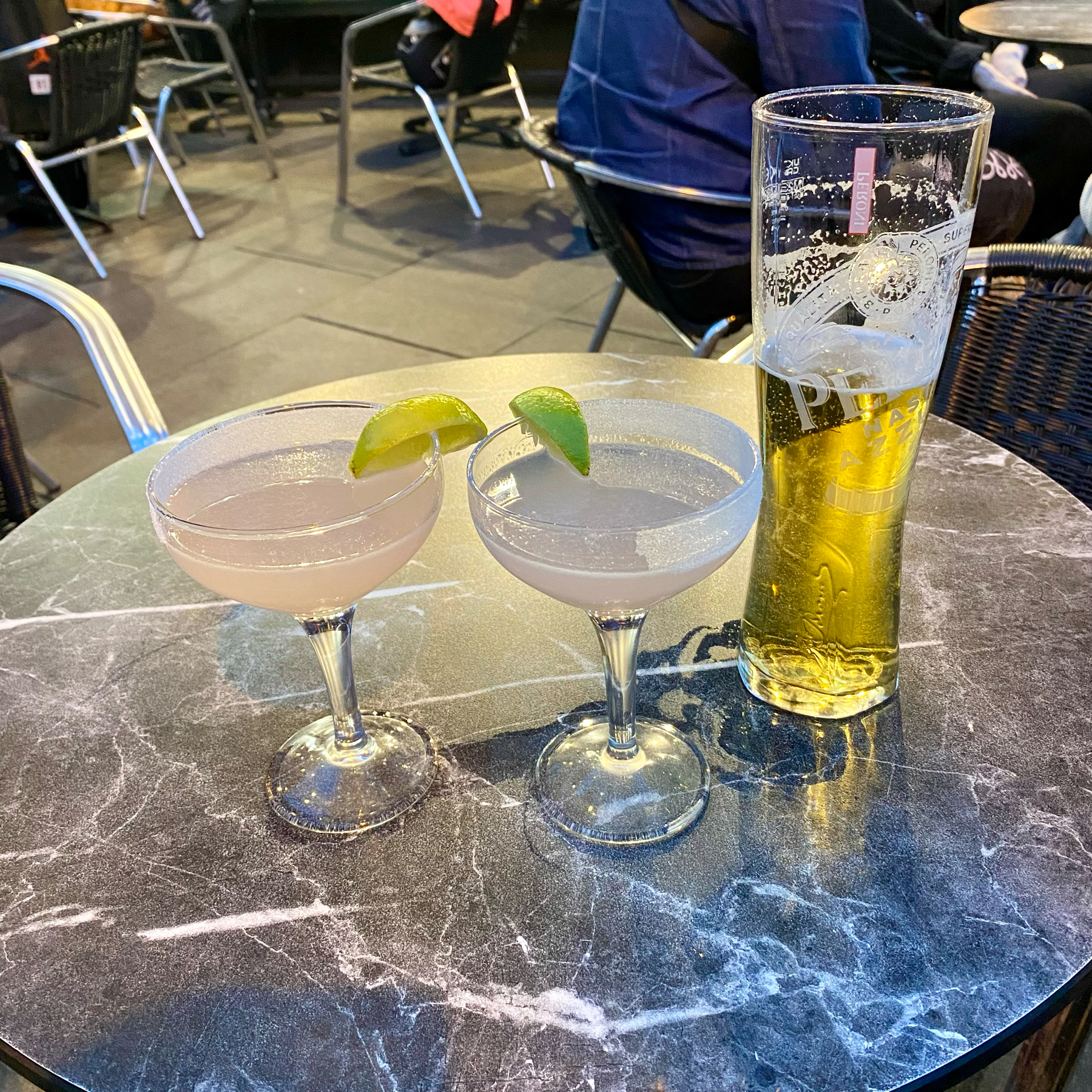 Two cocktails garnished with lime wedges and one tall glass of beer on a round, marble-top table in an outdoor seating area of a restaurant or bar.