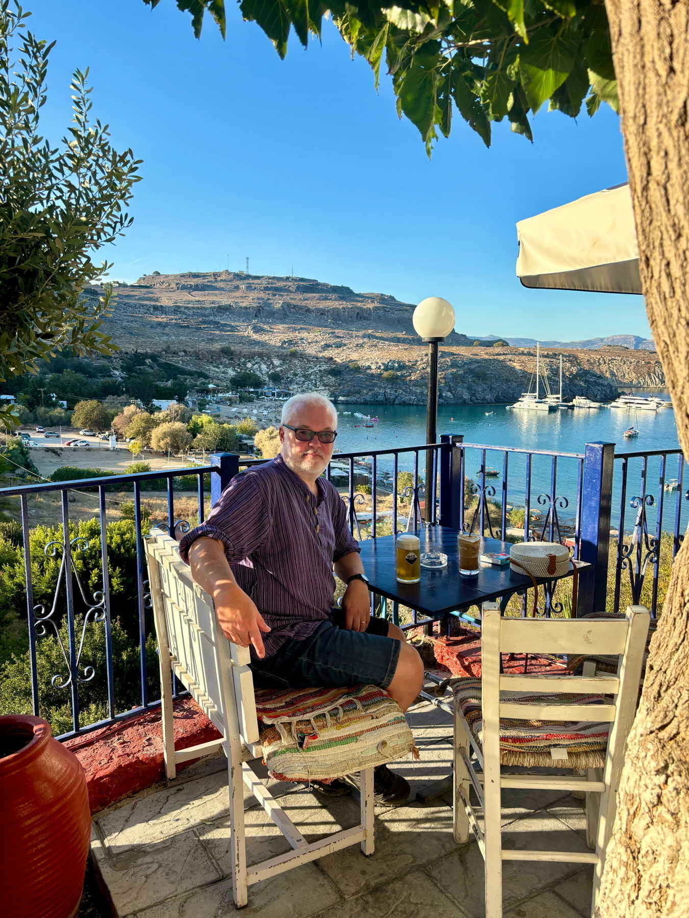 A man with glasses sits at a table on a balcony, enjoying a drink. The view overlooks a scenic bay with boats and a distant hillside. The setting is sunny and framed by a tree and railing.