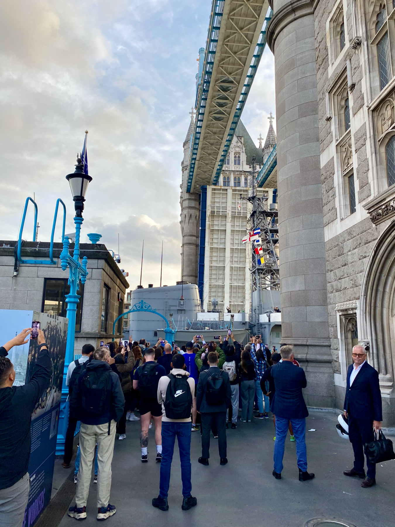 A group of people standing on the walkway near Tower Bridge in London, taking photos and looking up at the bridge. The image shows the bridge's structure, with its towering stone pillars and blue metal framework.