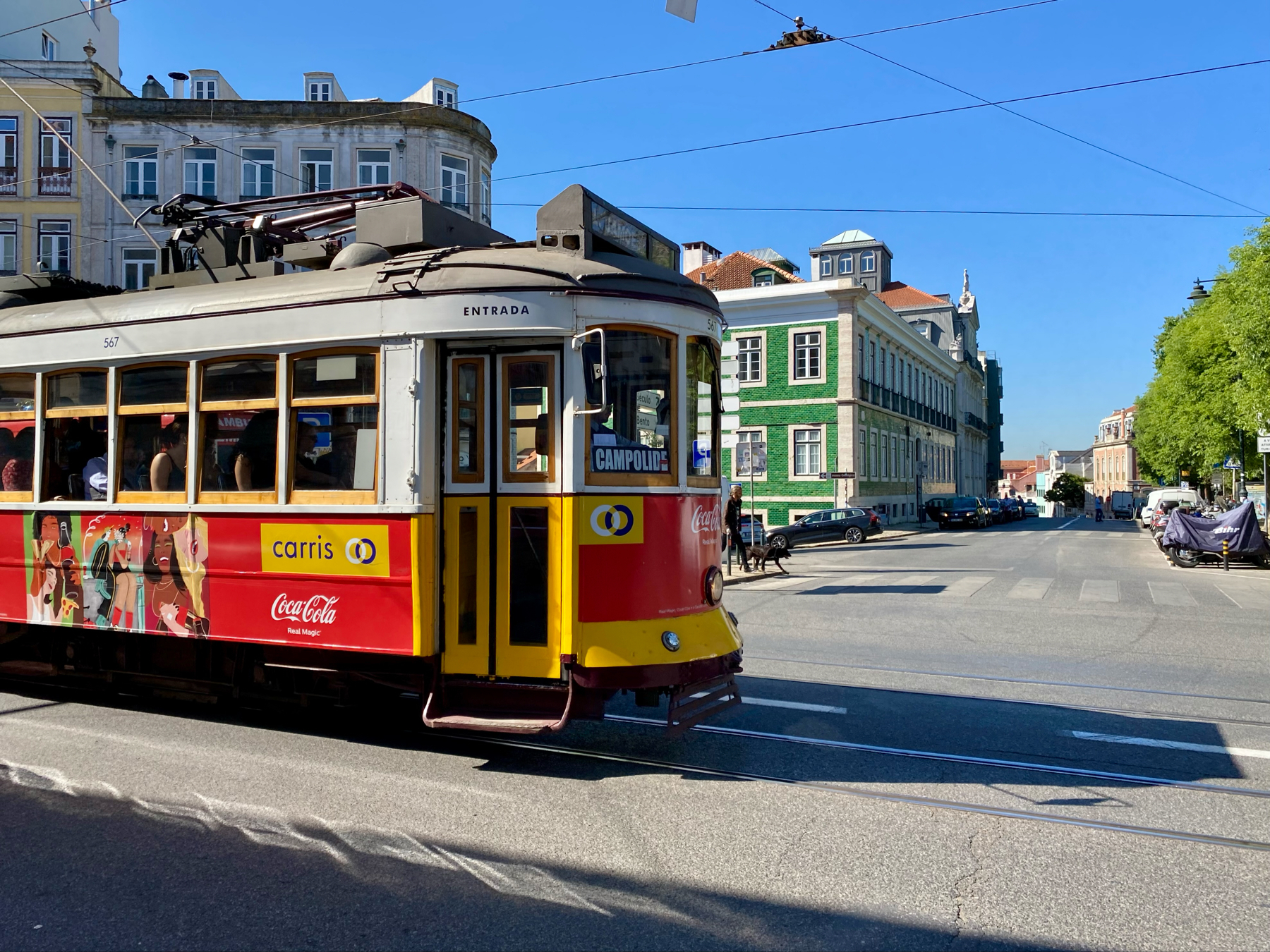 A vintage yellow and red tram with a Coca-Cola advertisement on its side travels on a street in a sunny urban setting with classic European architecture.
