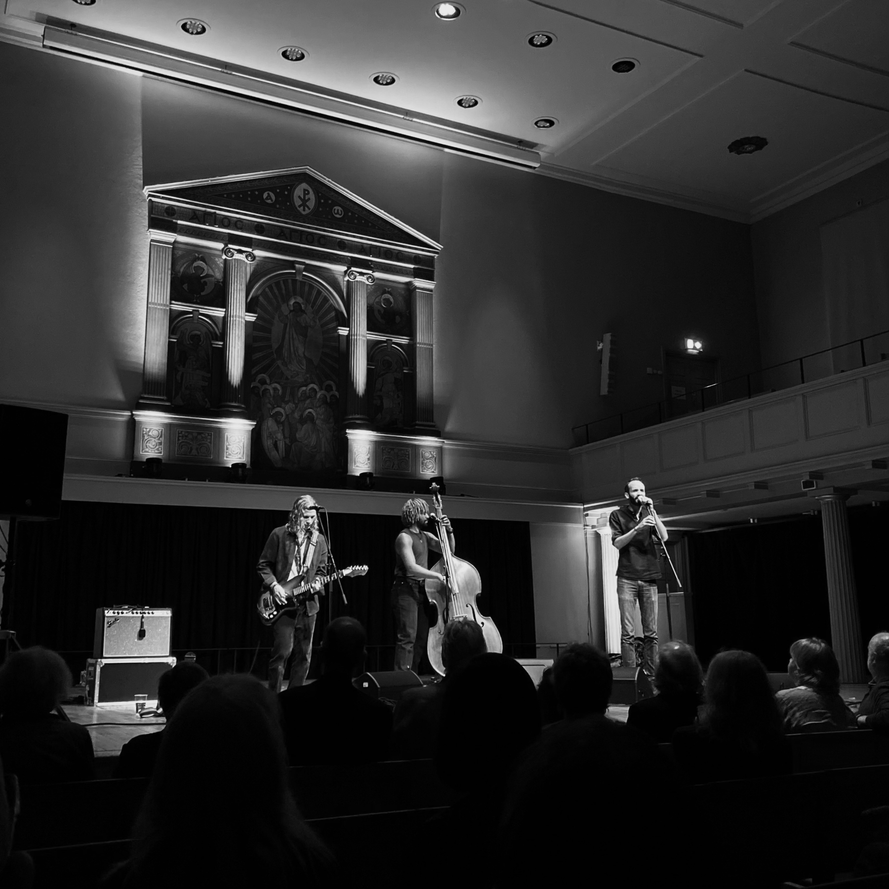 A black and white photo of a live music performance featuring three musicians. The setting includes an ornate stage backdrop with classical architectural details. The band consists of one person playing an electric guitar, another playing an upright bass, and a third singing 