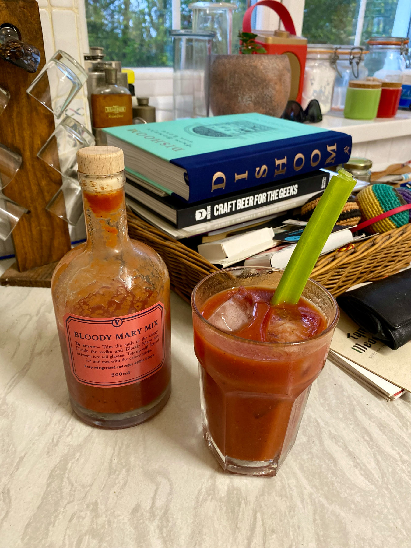 A Bloody Mary cocktail with a celery stick in a glass next to a bottle of Bloody Mary mix on a kitchen counter, with books and kitchen items in the background.