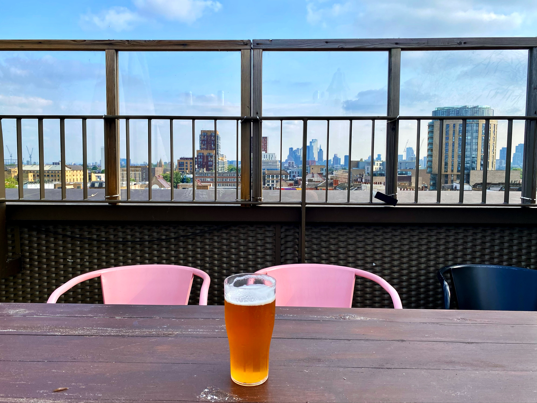 A rooftop scene with a wooden table, pink and black chairs, and a glass of beer in the center. The background features a metal railing and a cityscape under a blue, partly cloudy sky.
