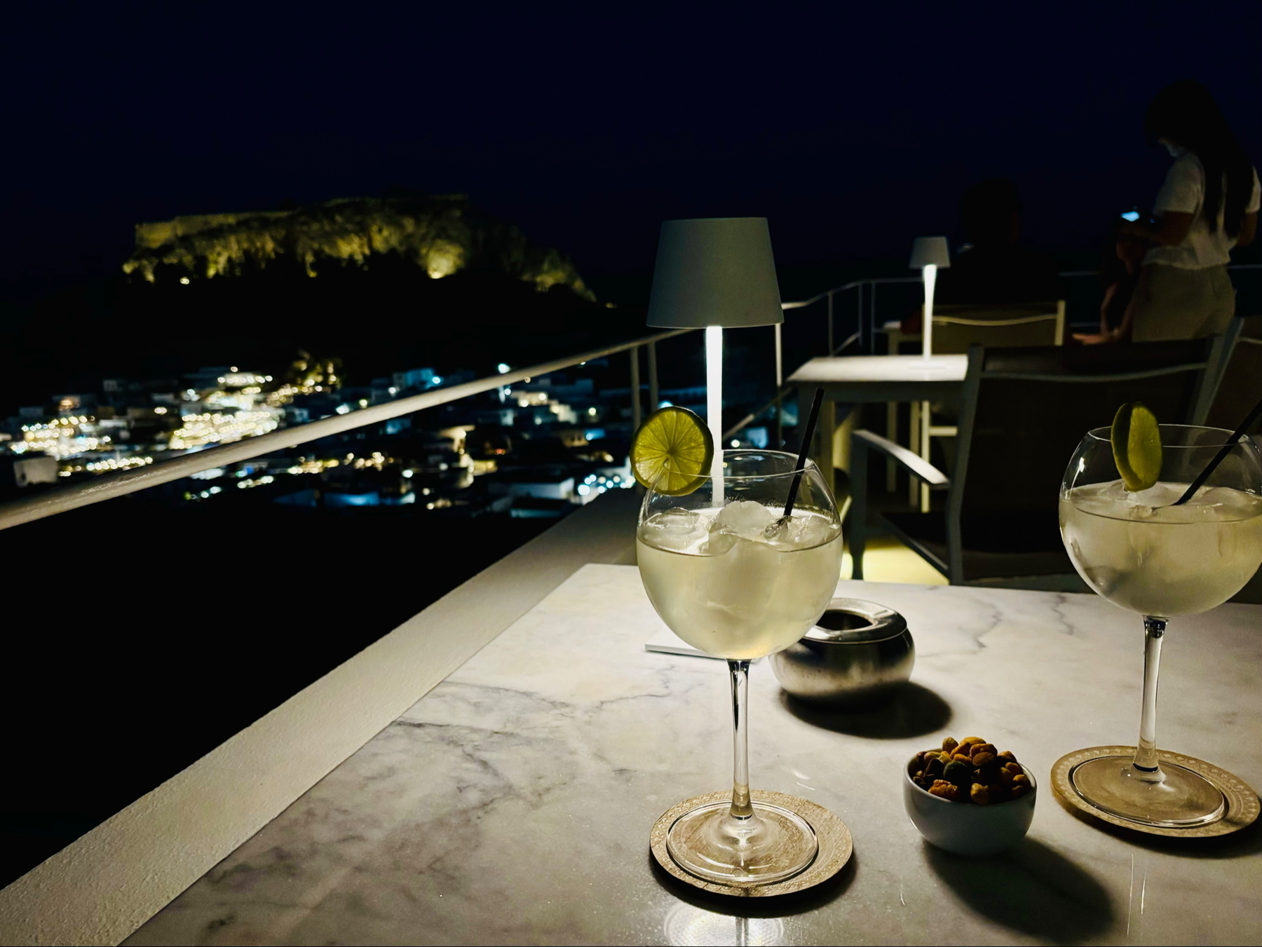 Two cocktails with lime and ice on a table at a rooftop bar, with a scenic view of a lit-up city and hill in the background. A small bowl of snacks is next to the drinks, and a couple of modern table lamps provide subtle lighting