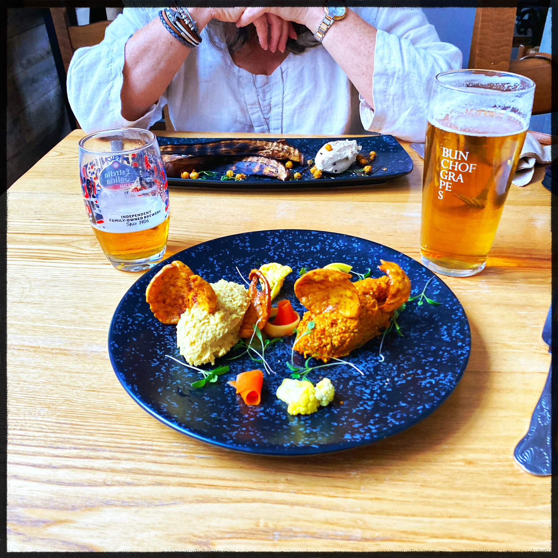 A dining setup featuring two plates of food on a wooden table with two glasses of beer. The plate in the foreground has a sophisticated presentation of what appears to be a dish with patés and garnished vegetables.