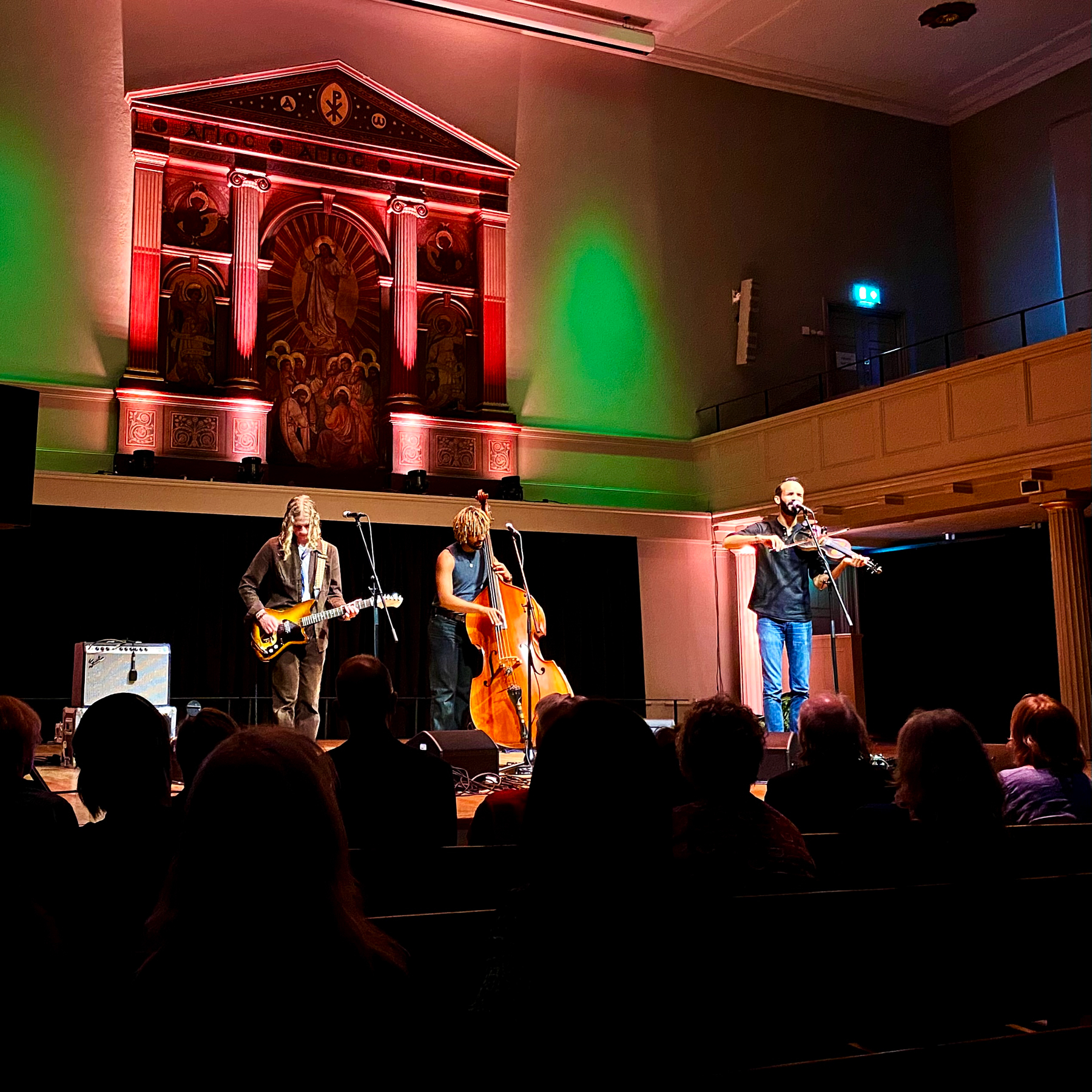 Three musicians perform on stage in a dimly lit venue. One plays an electric guitar, another stands by a double bass, and the third plays a violin. The backdrop features large, illuminated artwork and colorful lighting. An audience watches the performance.