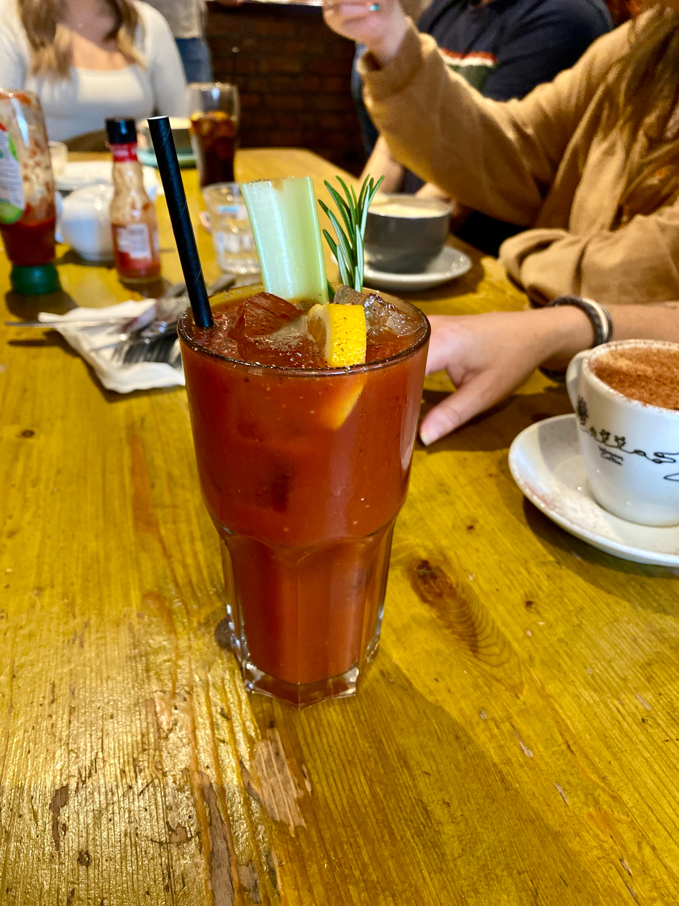 A Bloody Mary cocktail on a wooden table, garnished with celery, lemon and rosemary, with people and beverages in the background.