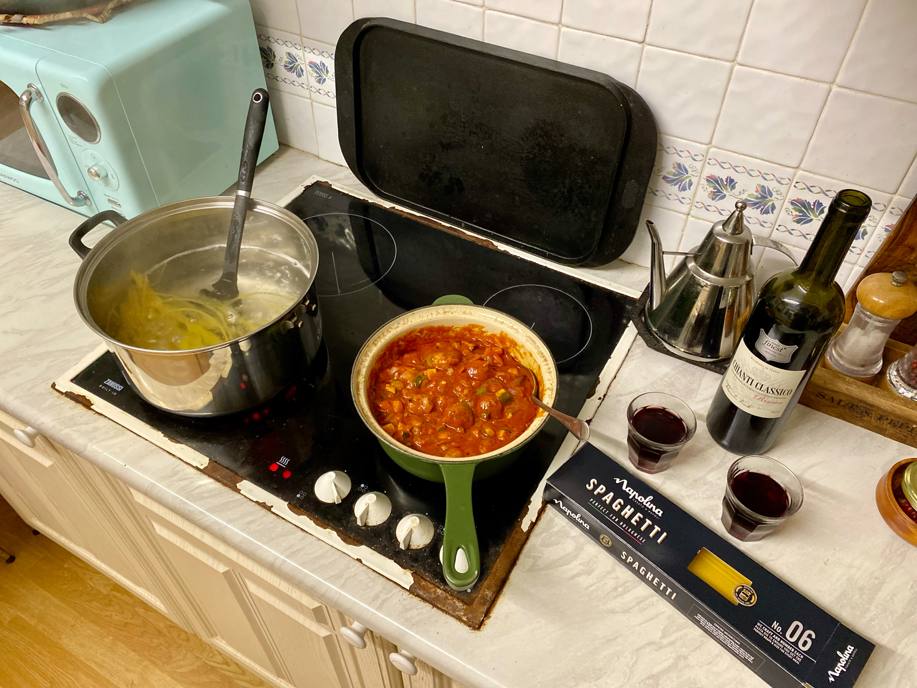 A kitchen countertop with a pot of boiling spaghetti, a pan of tomato sauce with meatballs and vegetables, two glasses of red wine, a wine bottle, a box of spaghetti, and various cooking utensils and condiments.