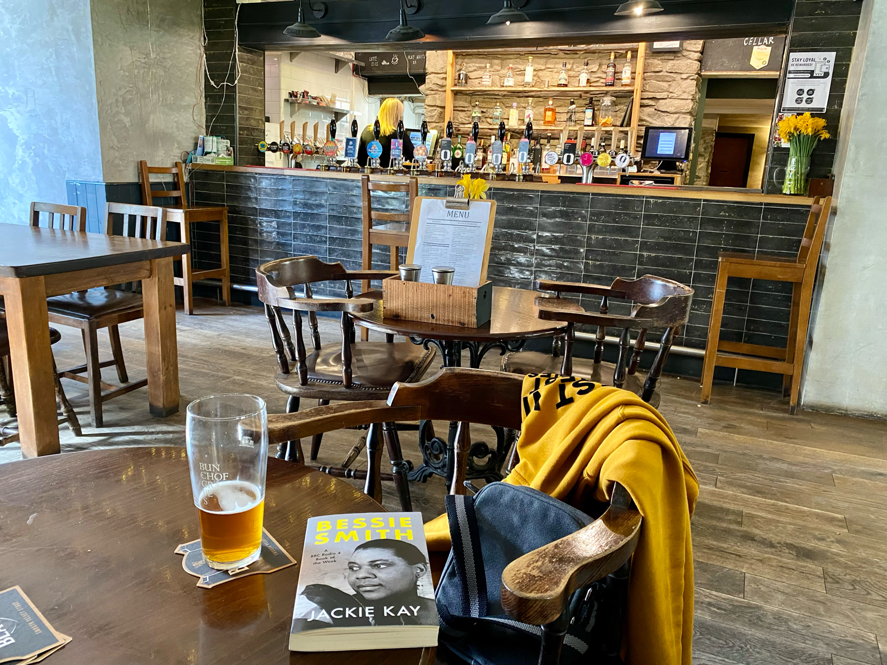 A pub interior with wooden tables and chairs, a half-full glass of beer on a table, and a book titled Bessie Smith by Jackie Kay. A bar with taps and bottles in the background, and a coat draped over a chair.